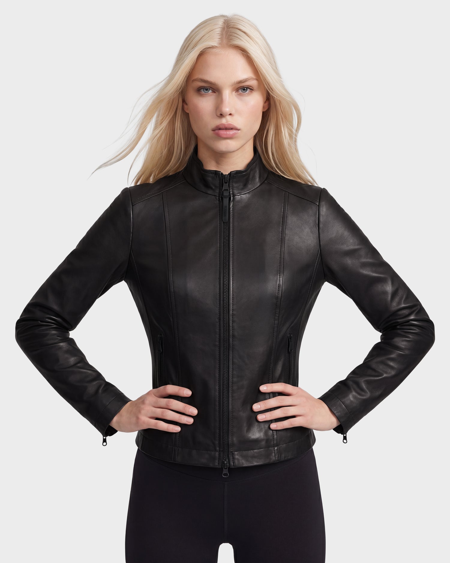 A woman with long blonde hair stands against a plain light background, wearing the Rudsak MARIAH - L124511 Black women’s leather jacket and black pants, her hands on her hips.