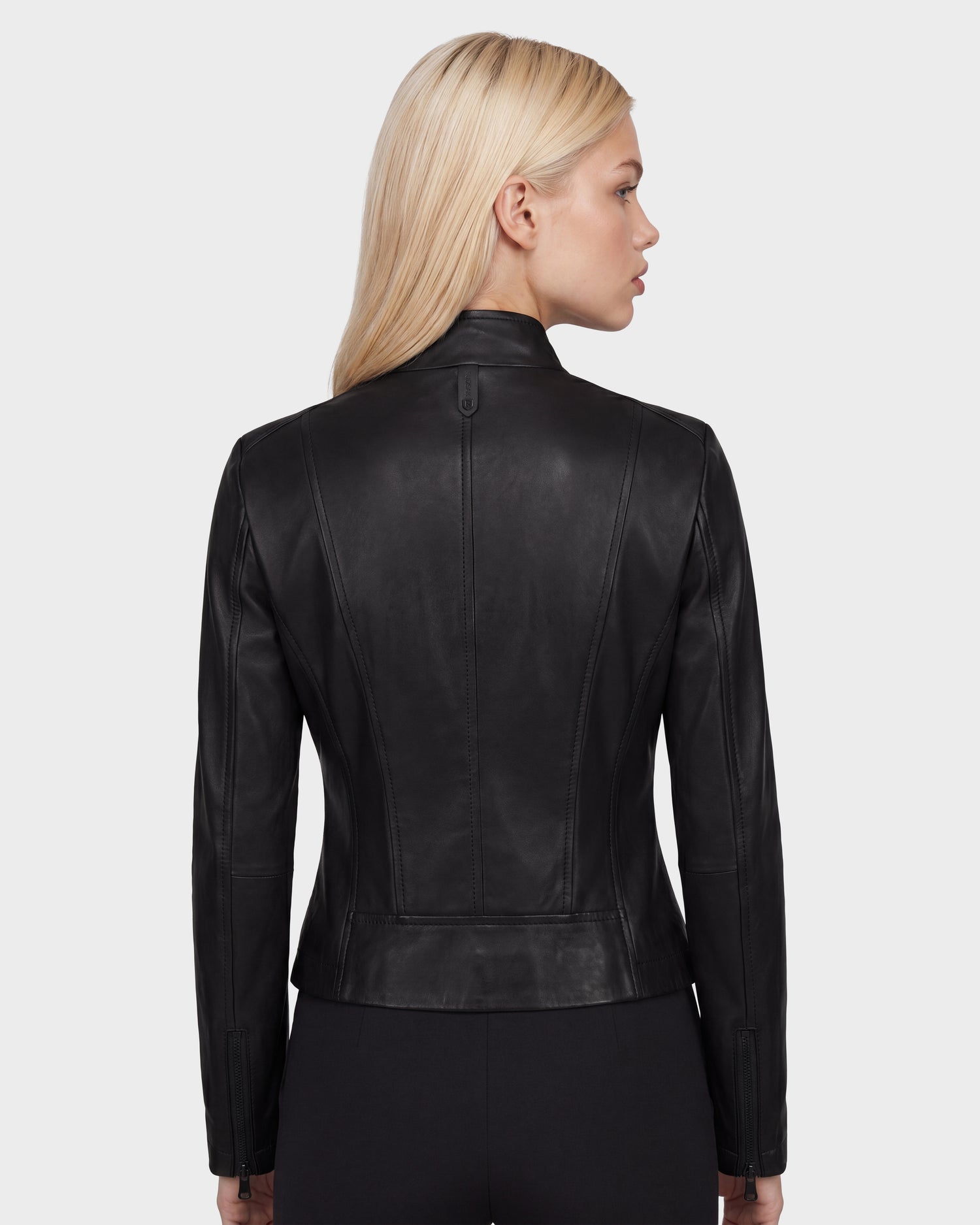A woman with long blonde hair wears the Rudsak MARIAH - L124511 Black fitted leather jacket and black pants, standing with her back to the camera against a plain light gray background.