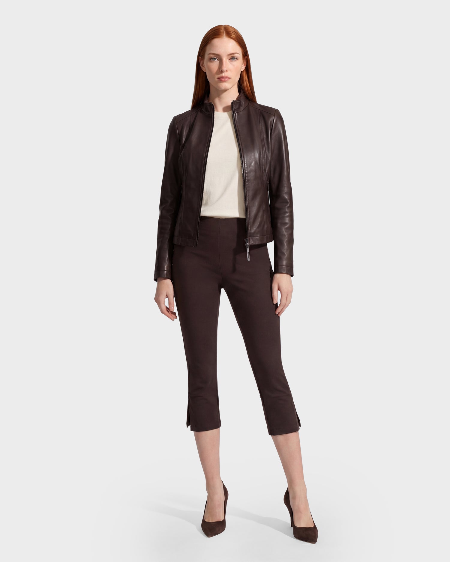 A woman with straight red hair wears the Rudsak MARIAH - L124511 Espresso women’s leather jacket over a white top, paired with black cropped pants and black heels, standing against a plain white background.