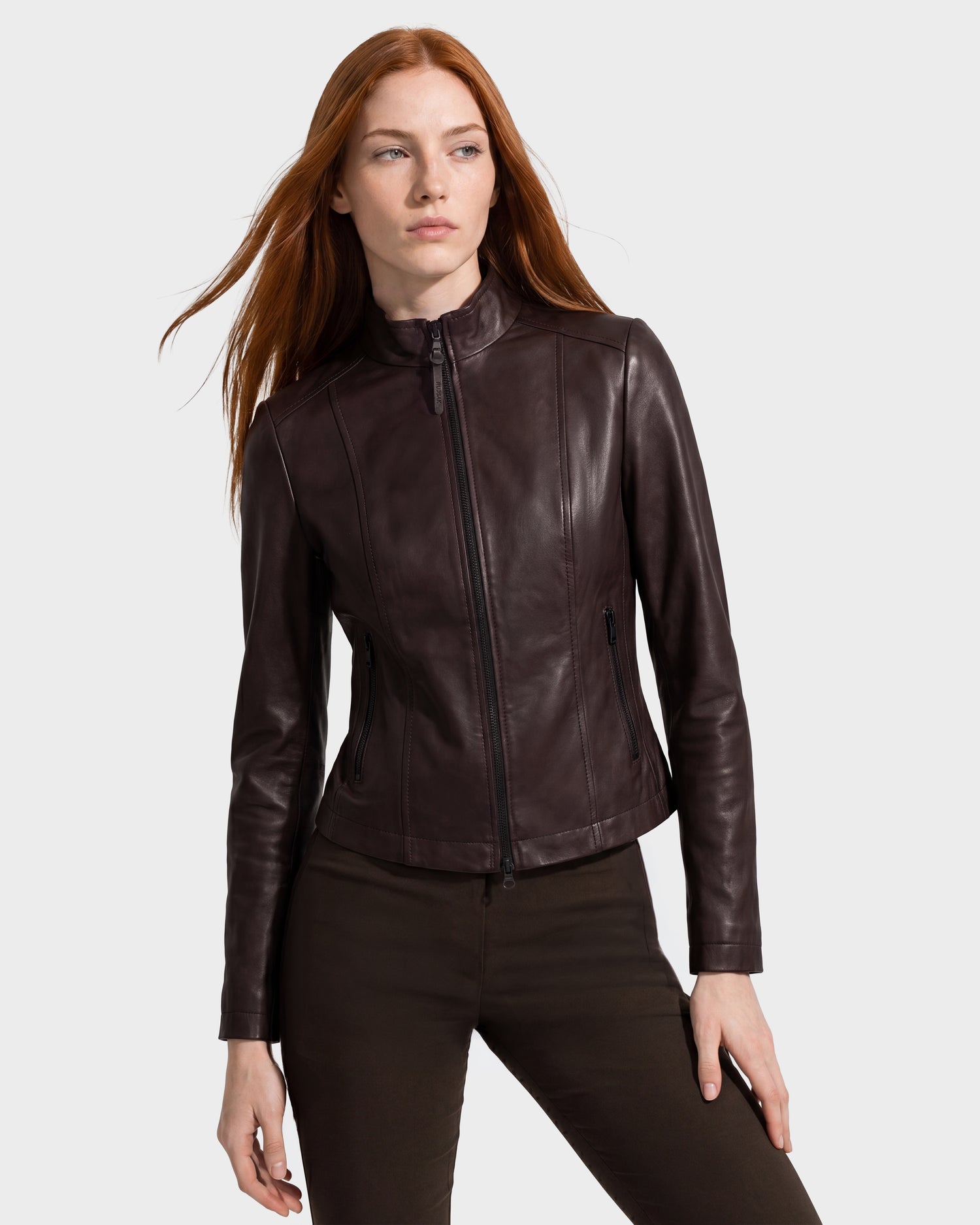 A woman with long red hair wears the Rudsak MARIAH - L124511 Espresso women’s leather jacket and dark brown pants, standing against a plain light gray background.