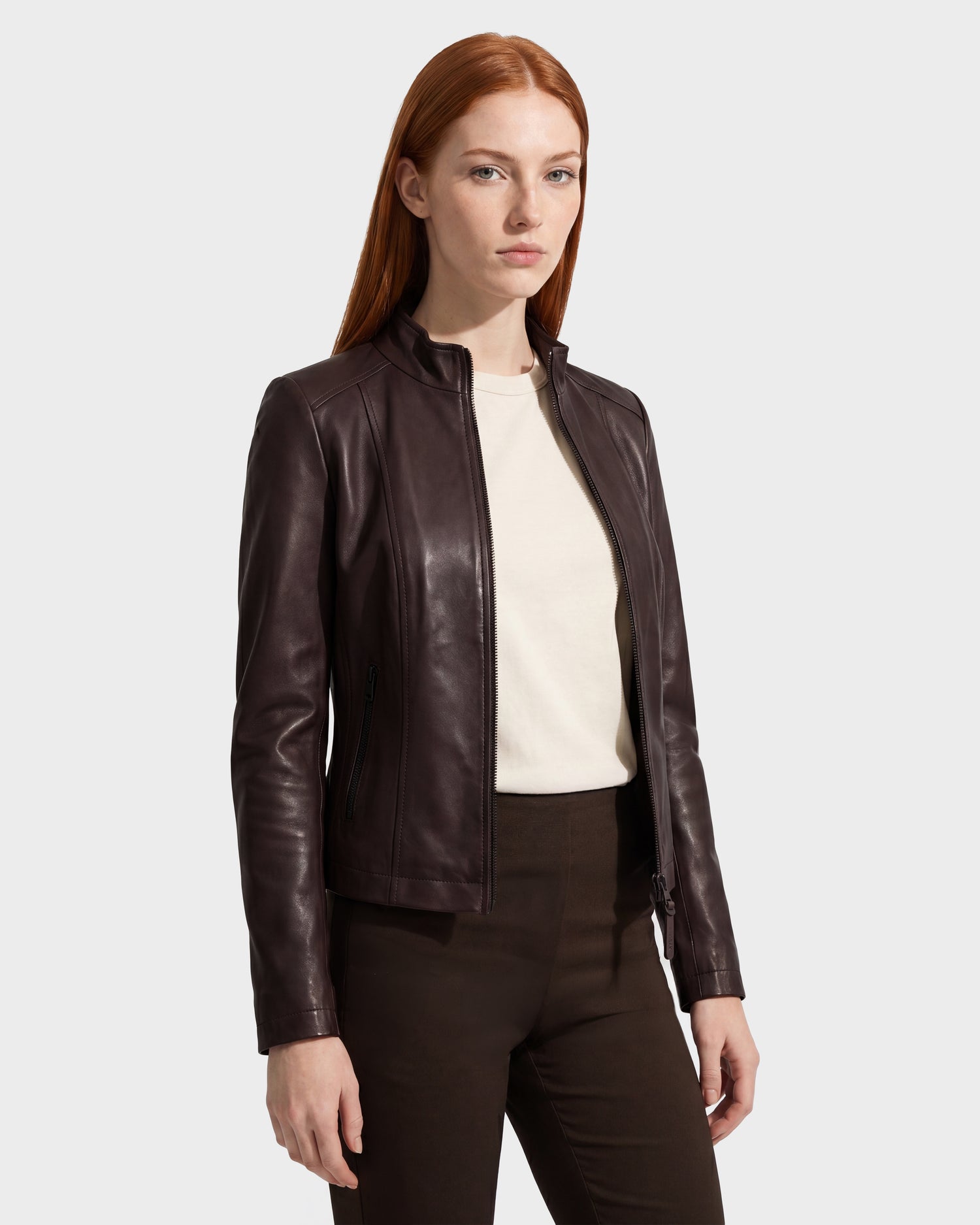 A woman with long red hair wears the Rudsak MARIAH - L124511 Espresso fitted women’s leather jacket over a white shirt and dark pants, standing against a plain light gray background.