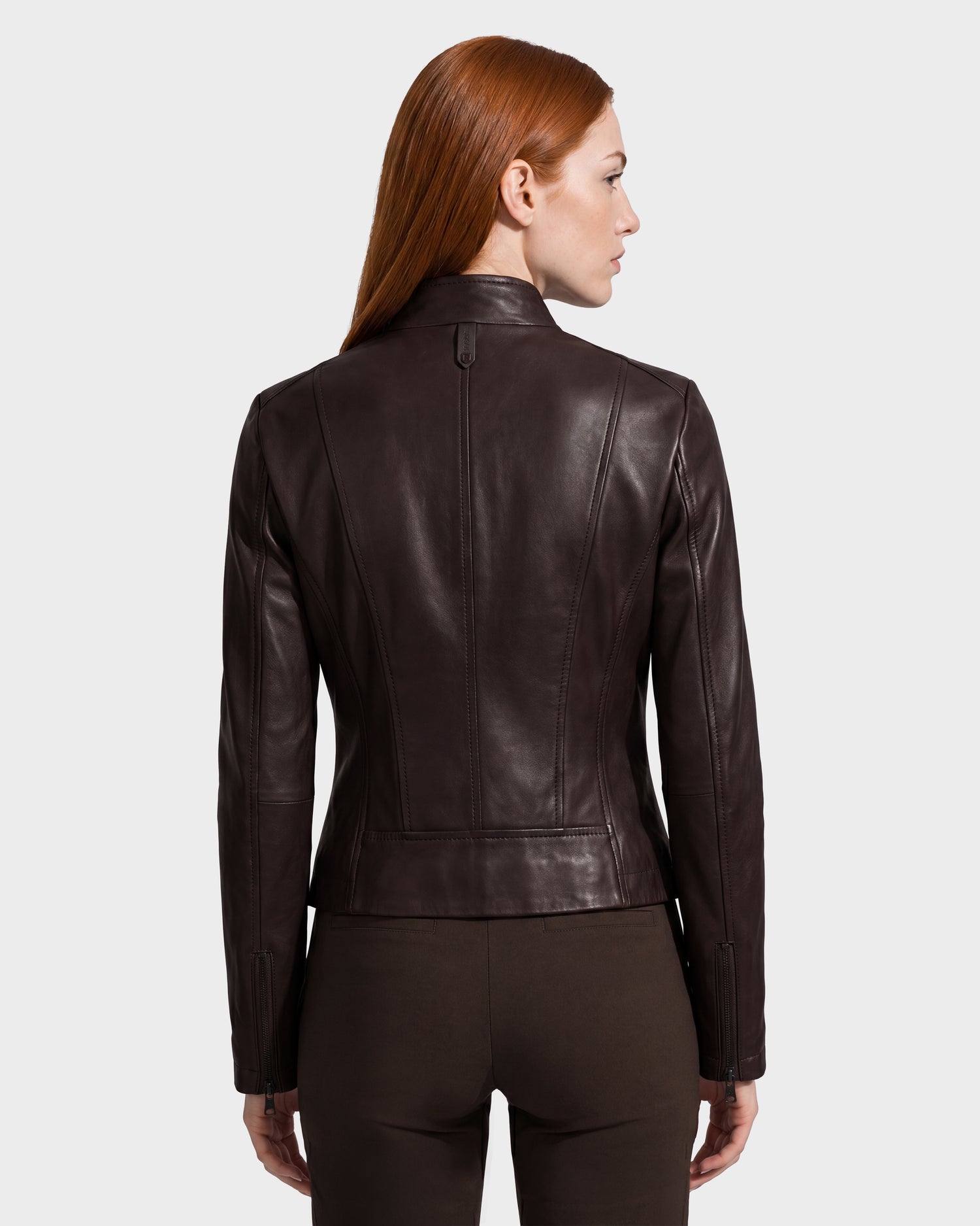 A woman with long red hair, shown from the back against a plain light background, wears the Rudsak MARIAH - L124511 Espresso fitted leather jacket paired with brown pants.