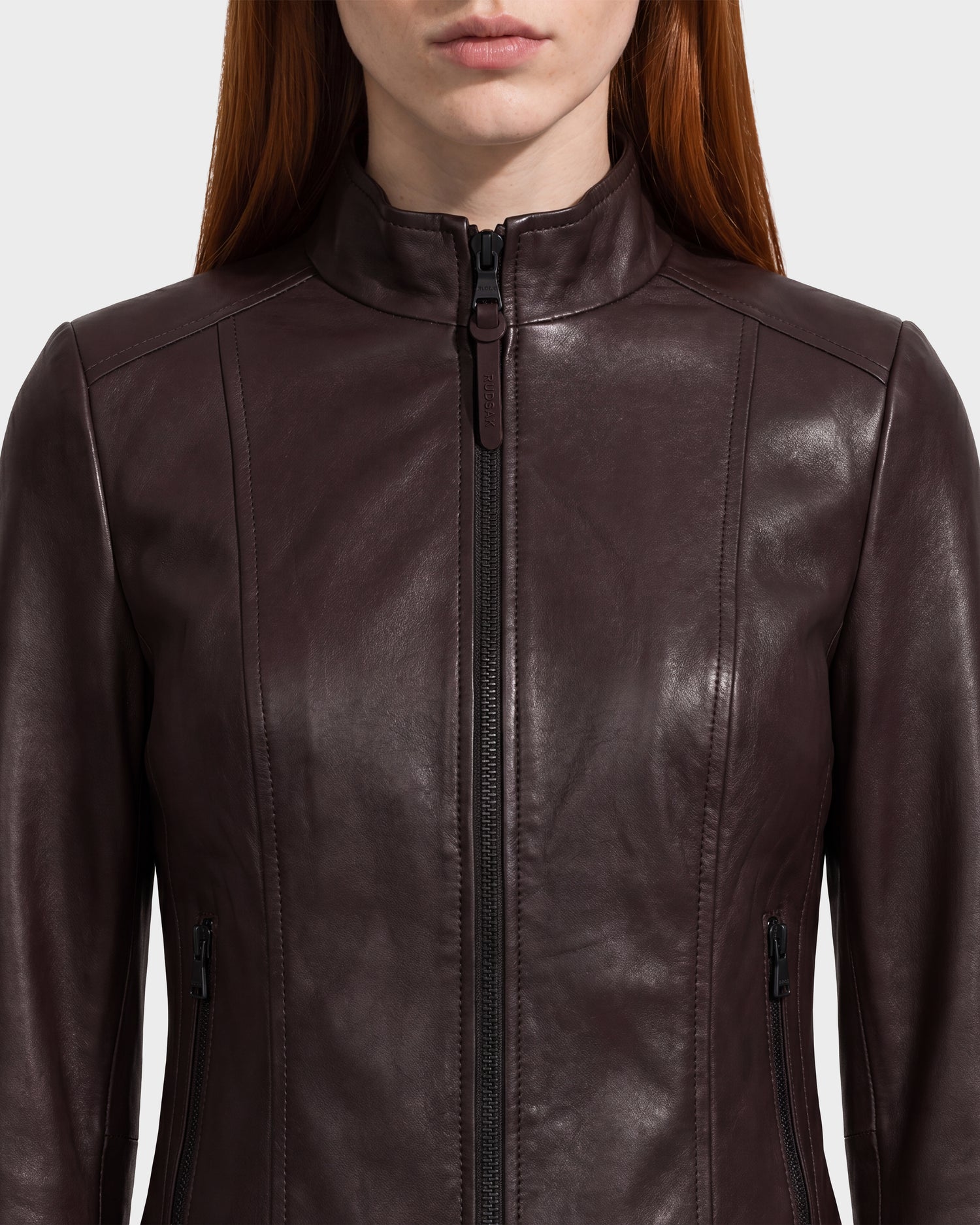 A woman wearing the Rudsak MARIAH - L124511 Espresso fitted women’s leather jacket, featuring a zippered front and high collar, shown from shoulders to mid-chest against a plain background.