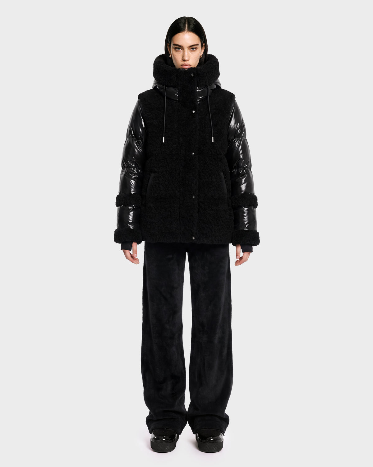 A person stands facing forward against a plain light gray background, wearing the Rudsak MARISSA - 8126728 Black down jacket featuring shiny sleeves and a fuzzy, winter-inspired texture, paired with loose black pants and black shoes.
