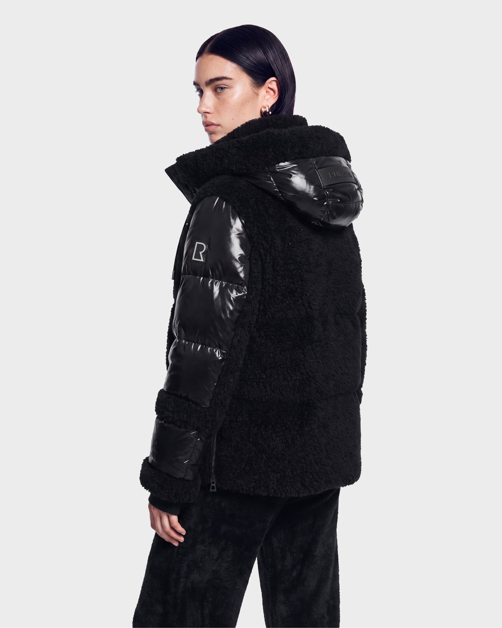 A person with dark hair is shown from the back, standing against a plain light background and looking over their shoulder while wearing the Rudsak MARISSA - 8126728 Black, a textured and shiny hooded down jacket.