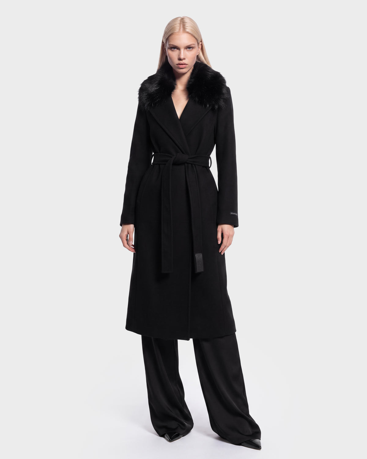 A woman wears the Rudsak MEL - 8126525 Black long coat, featuring a fur collar and waist tie, layered over black wide-leg pants and standing against a plain white background. This stylish coat is available for pre-order and is expected to ship by September 15th.
