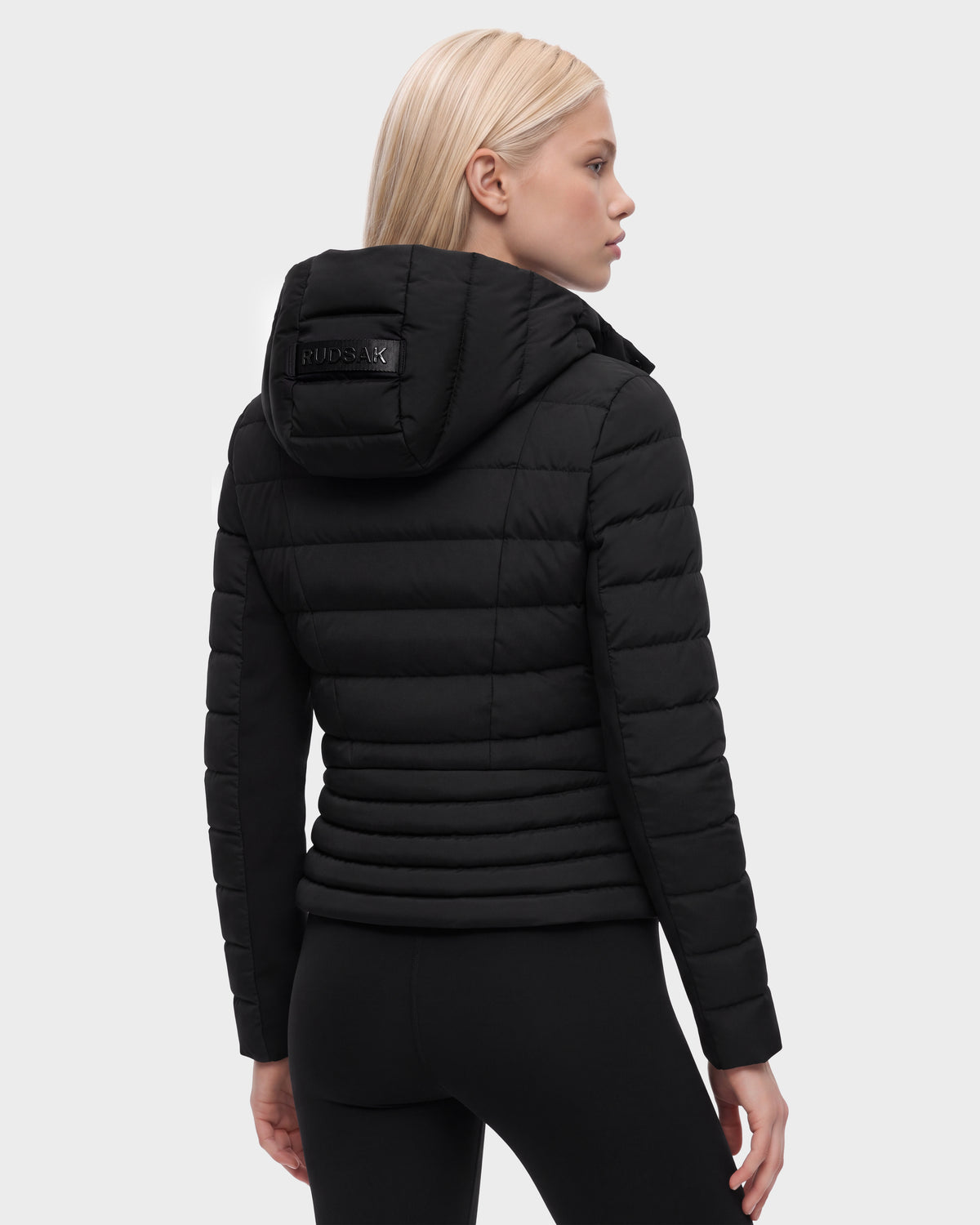 A person with straight blond hair is shown from the back, standing against a plain light gray background, wearing the Rudsak MELISSA - 8125054 Black women’s fitted puffer jacket with a hood and matching black pants. The jacket is insulated with RDS certified down for responsibly sourced warmth.