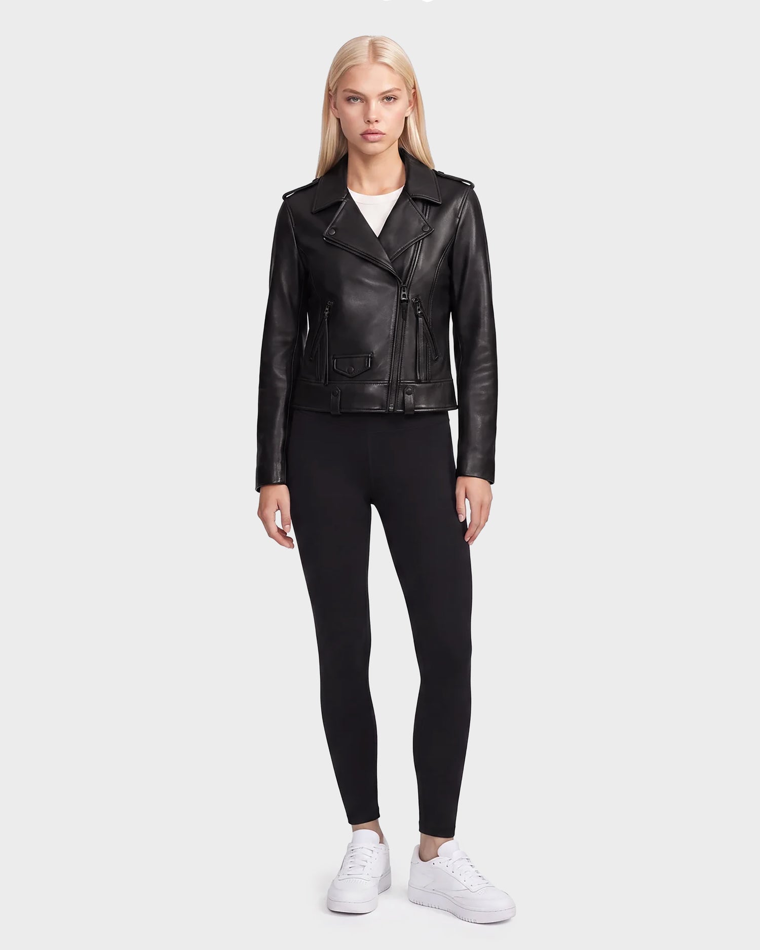 A woman stands facing forward against a plain light gray background, wearing the Rudsak MERGO - 8122009 BLACK lambskin leather biker jacket paired with black leggings, a white top, and white sneakers.