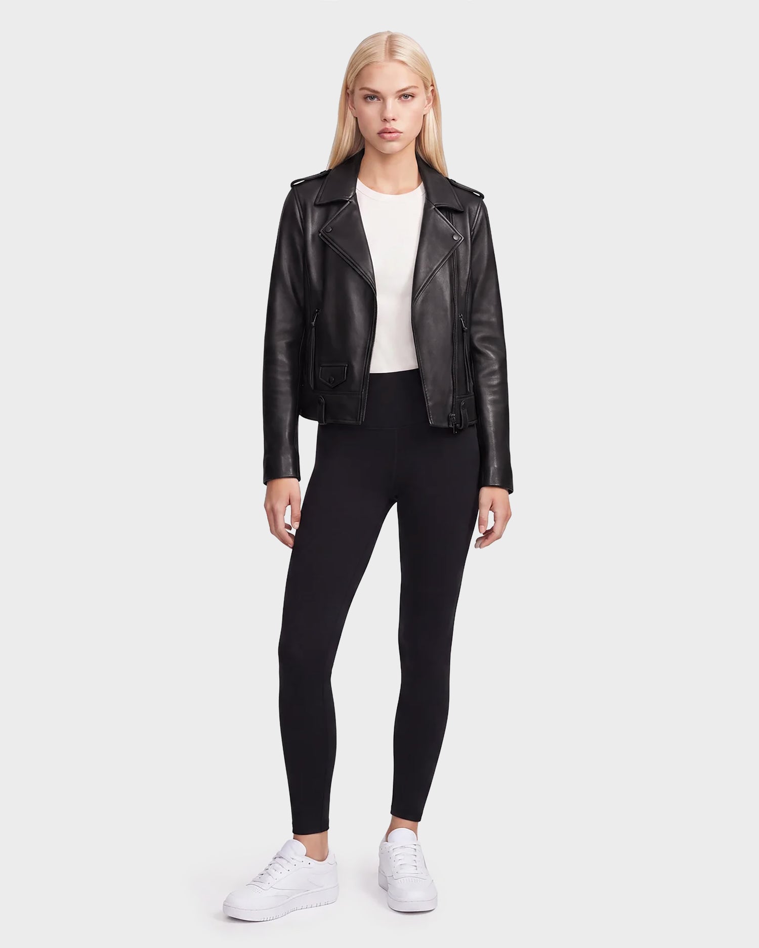 A woman stands facing forward wearing the Rudsak MERGO - 8122009 BLACK women’s black lambskin leather biker jacket, paired with a white shirt, black leggings, and white sneakers against a plain light background.