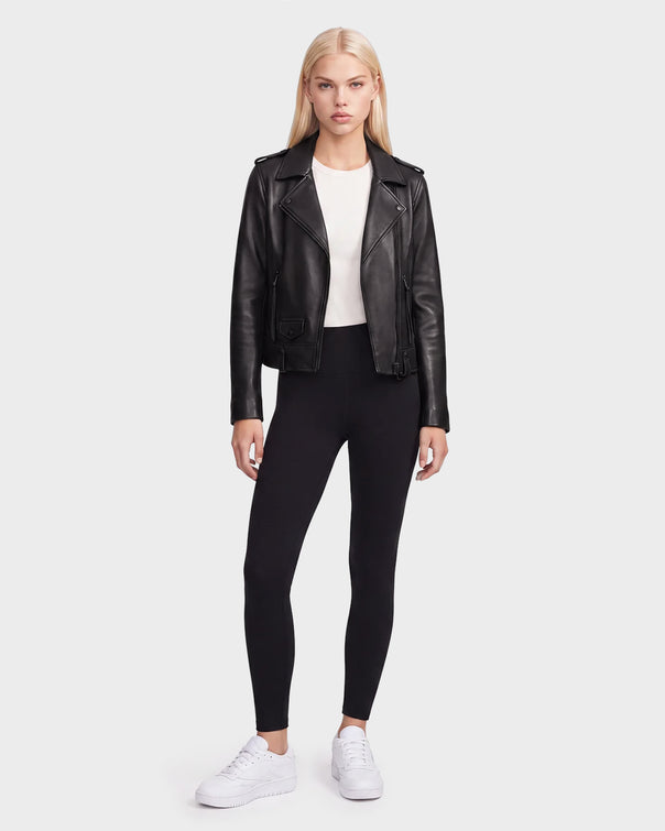 A woman stands facing forward wearing the Rudsak MERGO - 8122009 BLACK women’s black lambskin leather biker jacket, paired with a white shirt, black leggings, and white sneakers against a plain light background.