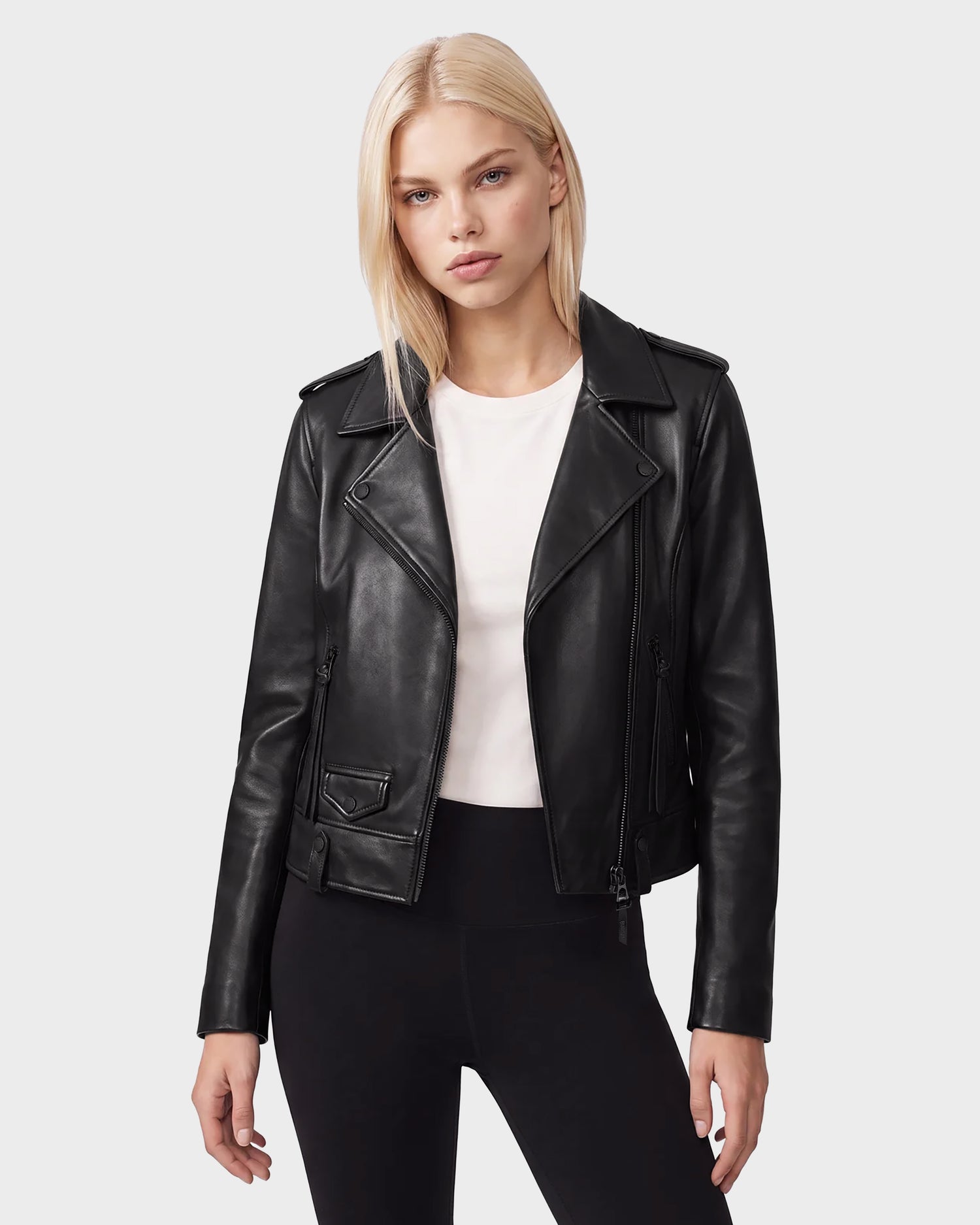 A woman with straight blonde hair wears the Rudsak MERGO - 8122009 BLACK lambskin leather biker jacket over a white shirt and black pants, standing against a plain light gray background.