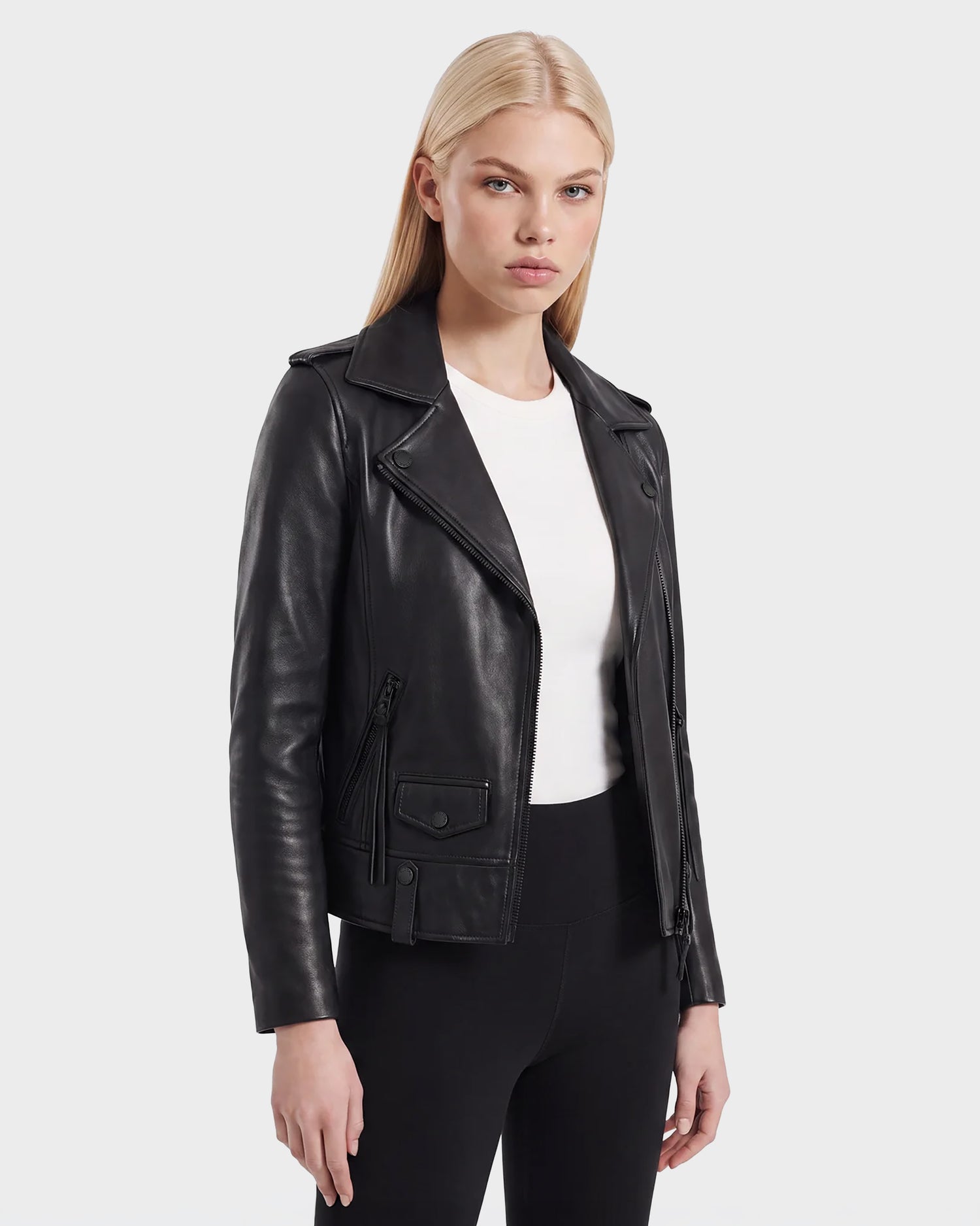 A woman with straight blonde hair wears the Rudsak MERGO - 8122009 BLACK lambskin leather biker jacket over a white shirt and black pants, standing against a plain light gray background.