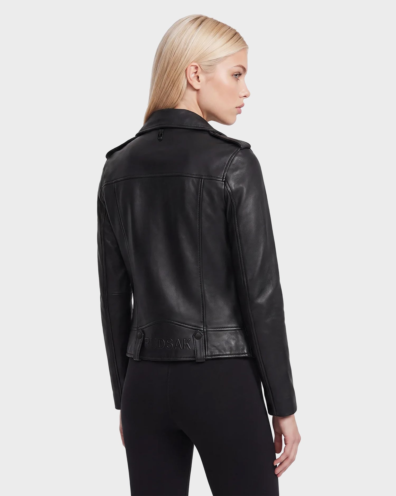 A woman with straight blonde hair wears the Rudsak MERGO - 8122009 BLACK fitted lambskin leather jacket and black pants, standing with her back turned and head slightly to the side against a plain light background.