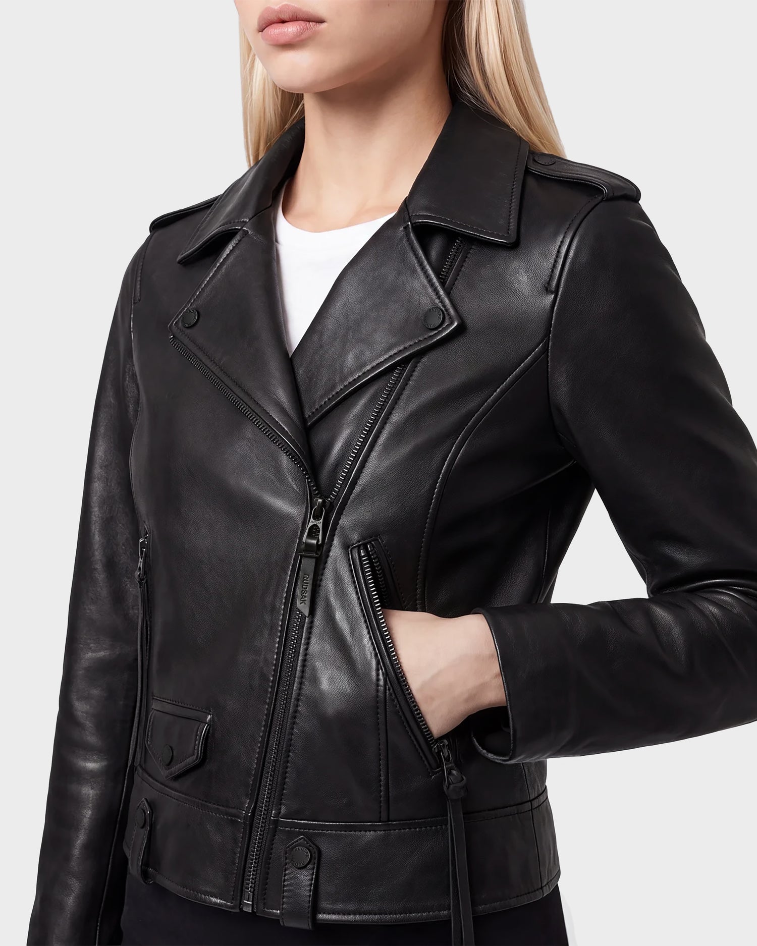 A woman is wearing the Rudsak MERGO - 8122009 BLACK fitted leather biker jacket, styled in black with a white shirt underneath. Her left hand rests in the zippered pocket of the women’s biker jacket, which features an asymmetrical front zip and collar. She has blonde hair down, and her face is partially visible.