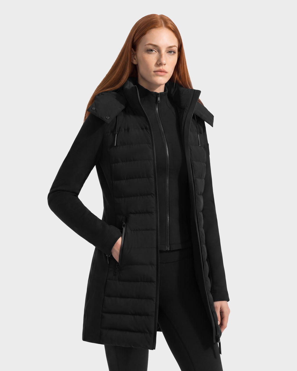A woman wears a black, hooded, knee-length quilted MERINA - 8125056 down vest over a matching long-sleeved transitional jacket.