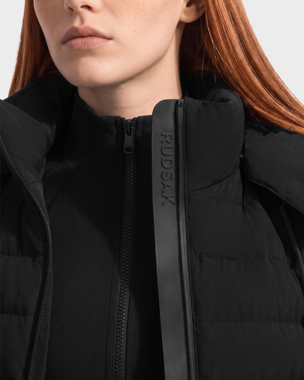 Close-up of a person wearing a black Rudsak MERINA - 8125056 jacket, a quilted down style with a high collar and a vertical zipper flap featuring embossed text.