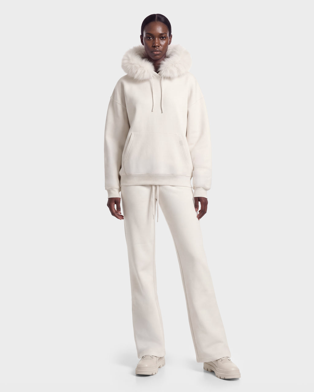A person wears the MILENA - 8526689 Vanilla by Rudsak—a plush off-white hoodie with faux-fur trim, matching pants, and chunky shoes—standing against a plain white background.