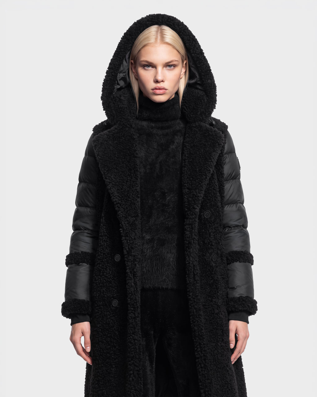 A woman with straight blond hair is shown standing against a plain white background, facing forward with a neutral expression, wearing the NOELLE - 8126971 Black long hooded coat by Rudsak. The coat features down insulation, a faux fur texture, and leather sleeves.