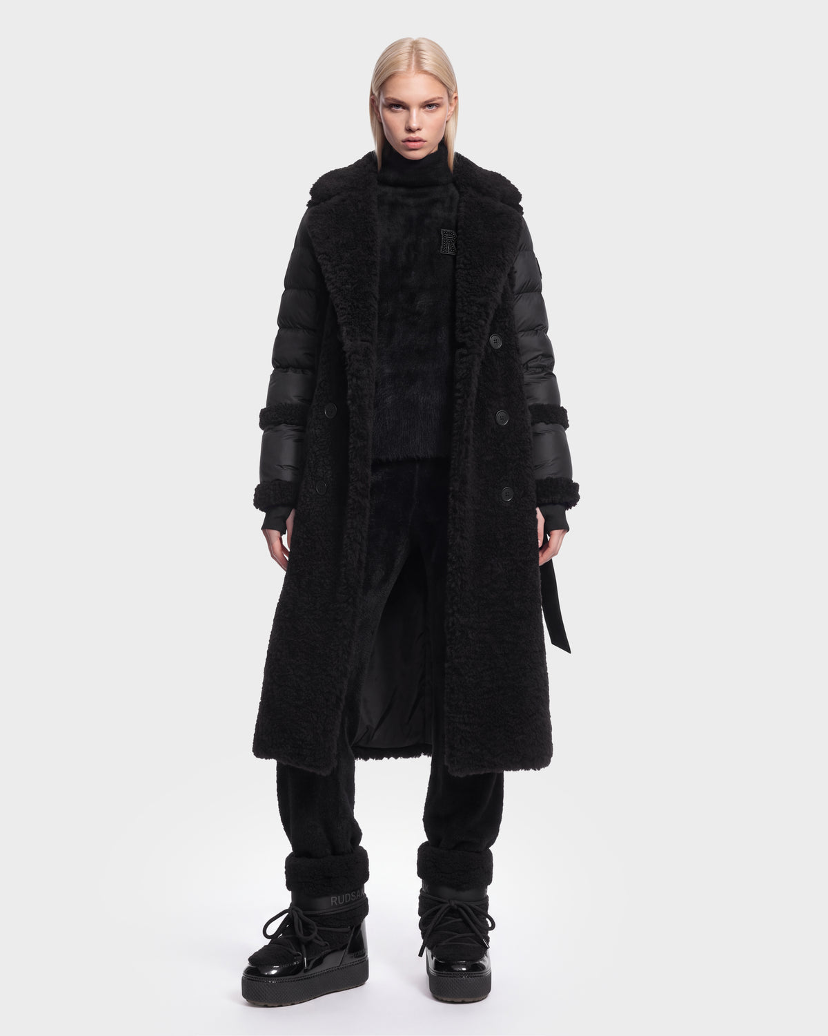 A person with straight blonde hair wears the Rudsak NOELLE - 8126971 Black long faux-fur coat with puffer sleeves, paired with black pants, black boots, and a high-neck top, standing against a plain white background.