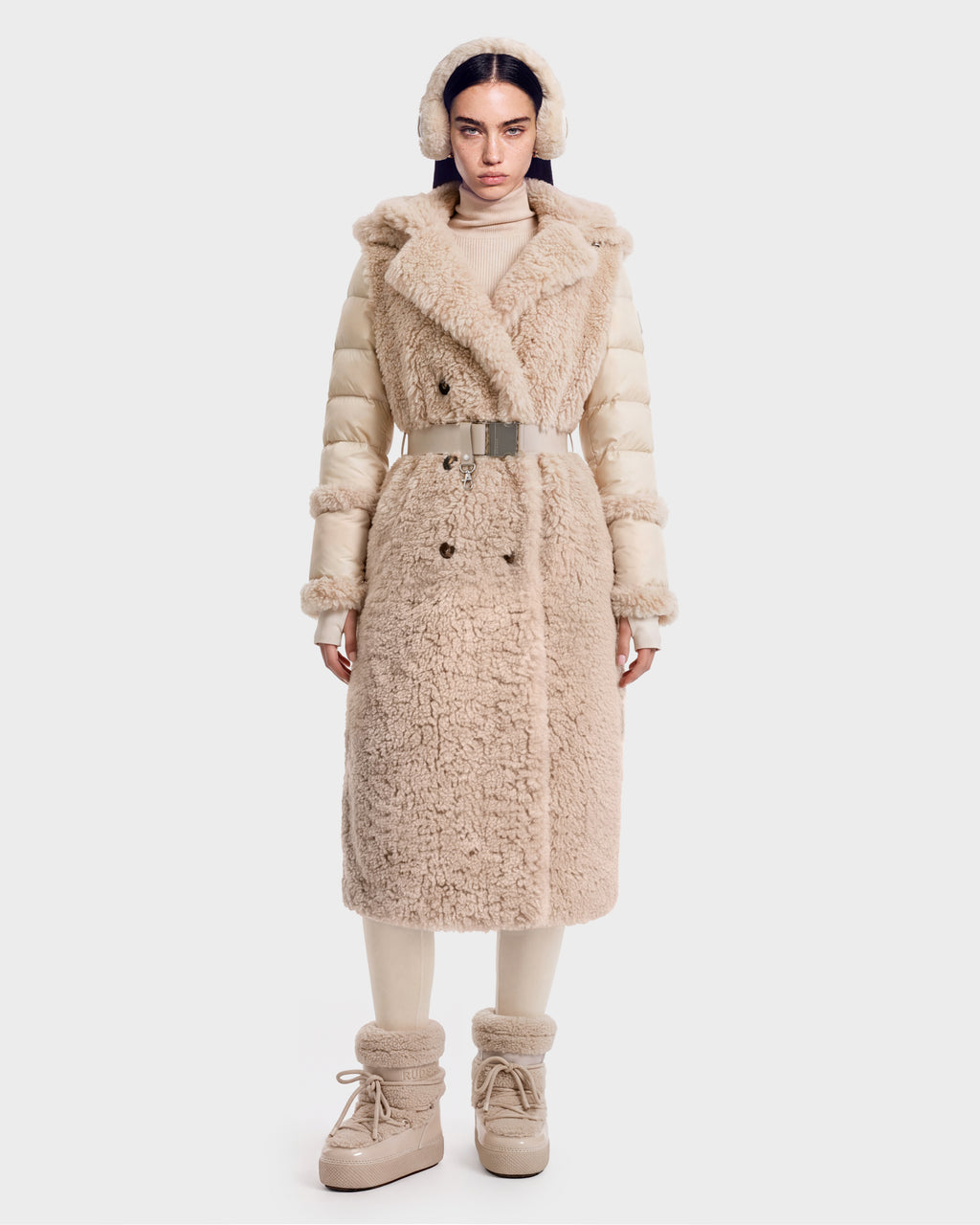 A woman stands facing forward, wearing the Rudsak NOELLE - 8126971 Cream plush faux fur coat with a belt, paired with matching ear muffs, boots, and a turtleneck. The outfit is cozy and winter-appropriate, featuring soft textures and neutral tones.