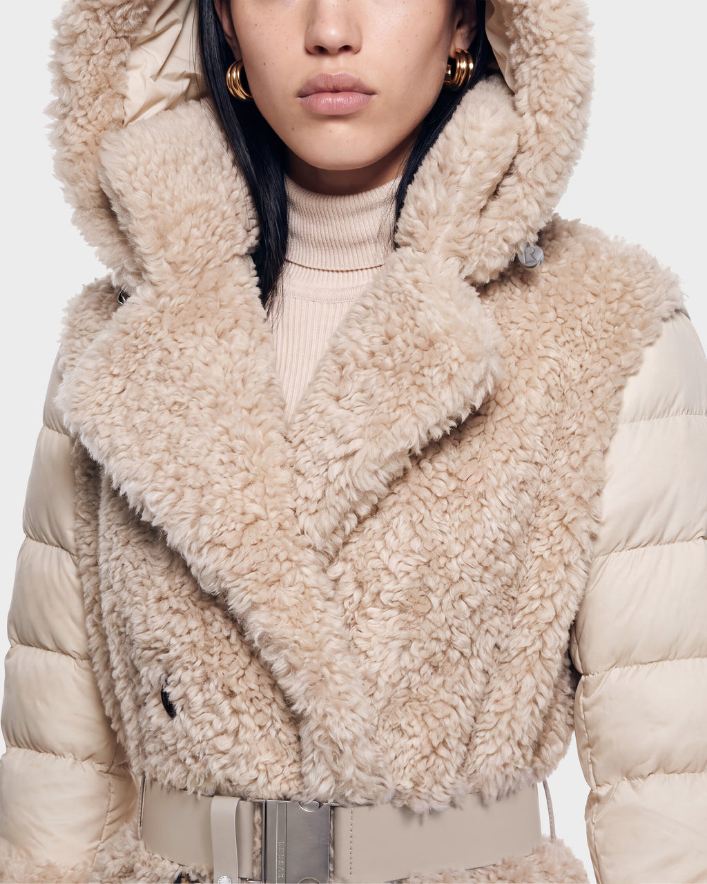 A woman wearing the Rudsak NOELLE - 8126971 Cream faux fur coat with a large hood, puffy sleeves, wide collar, turtleneck sweater, hoop earrings, and a wide belt. The photo is cropped to show her mid-face and upper torso.