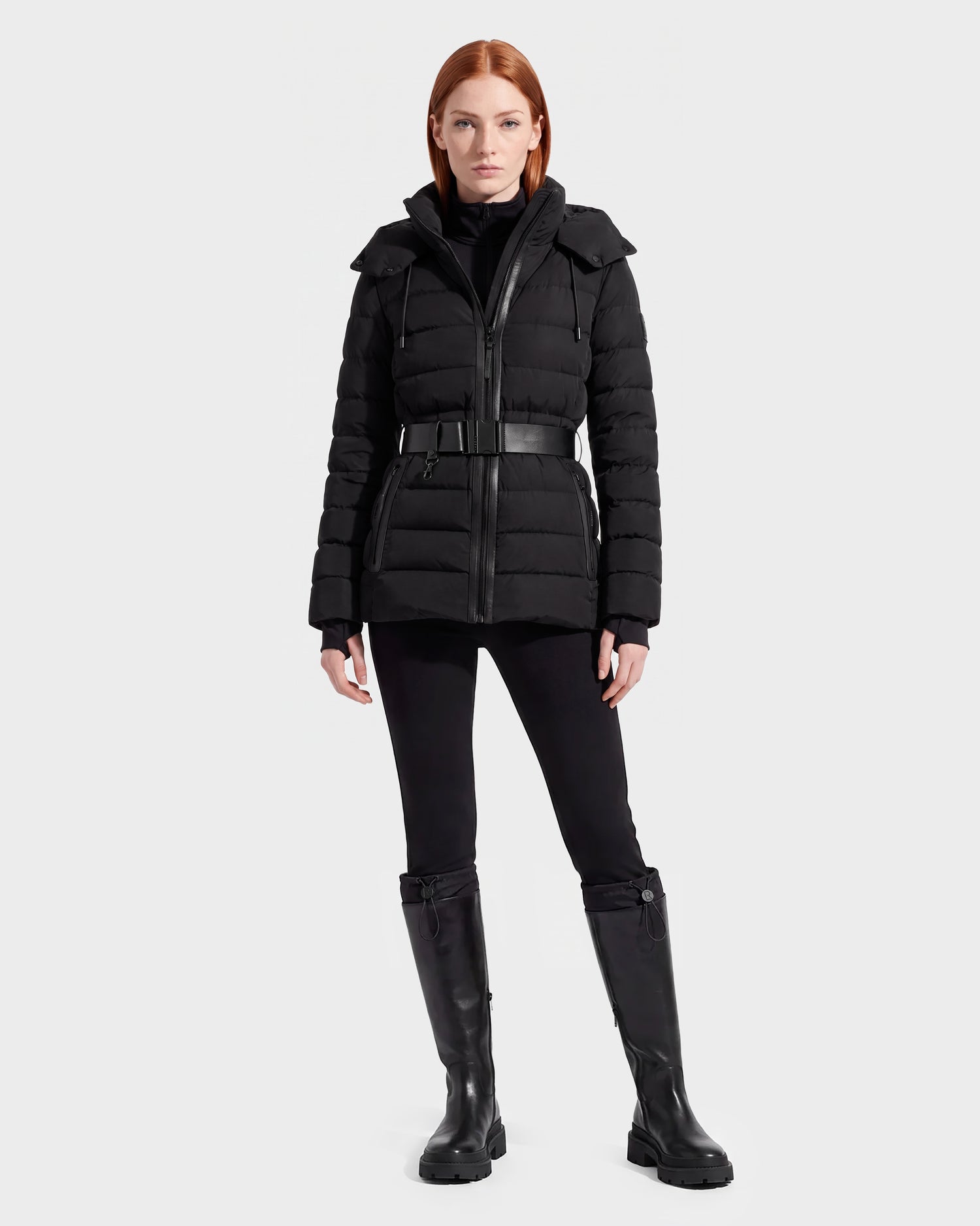 A woman stands facing forward against a plain white background, wearing the Rudsak PELIN S - 8124813 Black belted women's down puffer jacket, along with black pants and knee-high black boots.