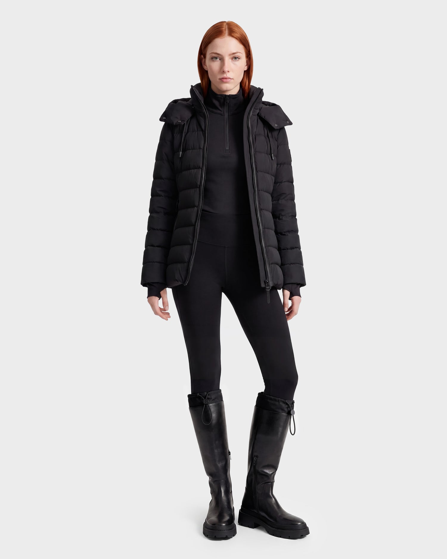 A woman with straight red hair, dressed in the Rudsak PELIN S - 8124813 Black women's down puffer jacket, a black top, black leggings, and black knee-high boots, stands facing forward against a plain white background.