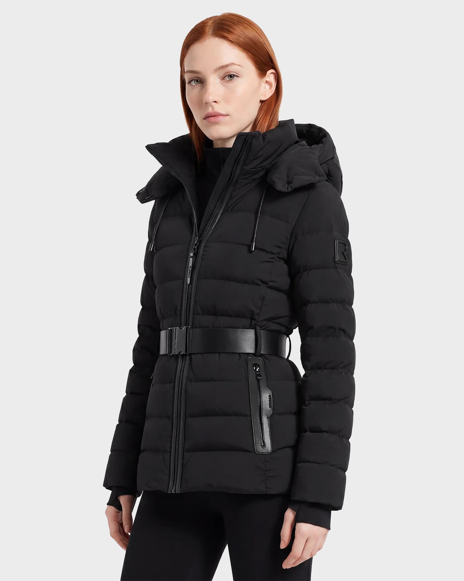 A woman with straight red hair wears the Rudsak PELIN S - 8124813 Black belted quilted puffer jacket with a hood and zippered pockets, standing against a plain light gray background.