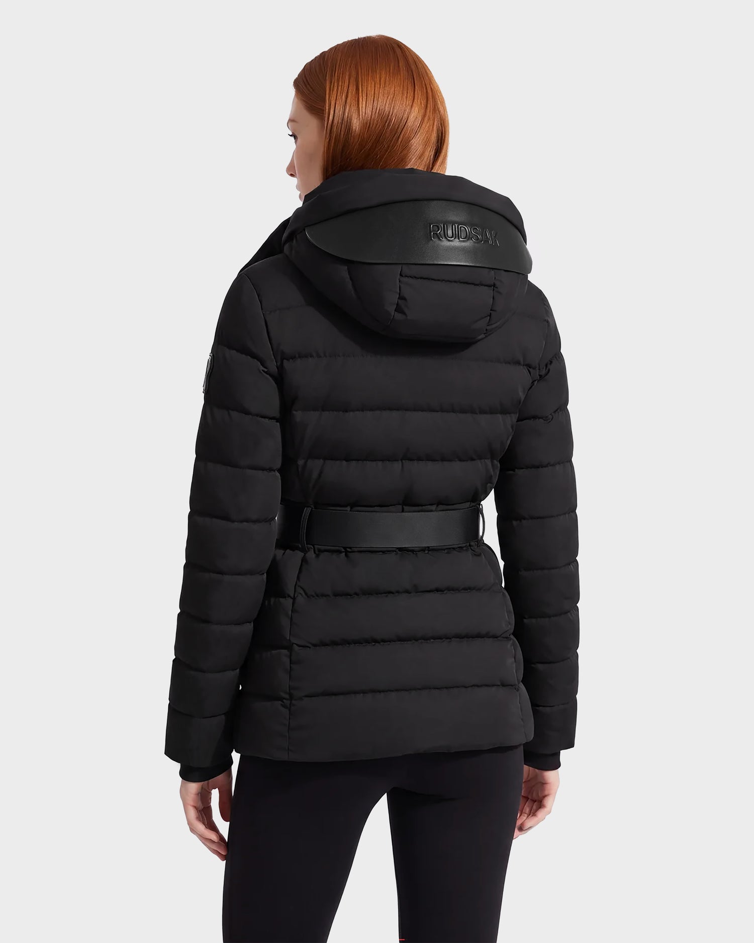 A woman with straight red hair is shown from the back against a plain light background, wearing the Rudsak PELIN S - 8124813 Black quilted puffer jacket, featuring a hood and belt.