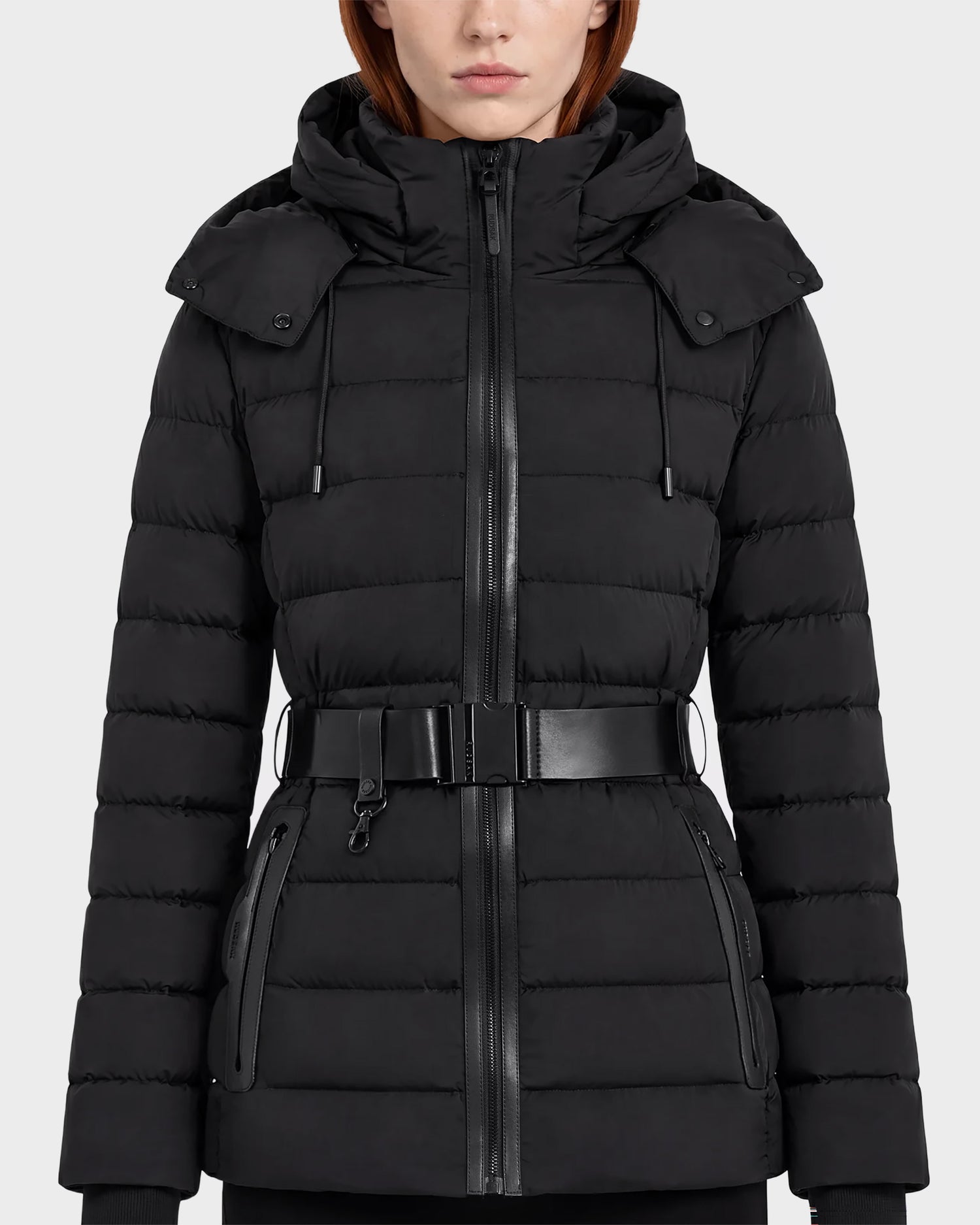A woman wearing the Rudsak PELIN S - 8124813 Black belted quilted puffer jacket with a hood and zippered pockets, standing against a plain light gray background.