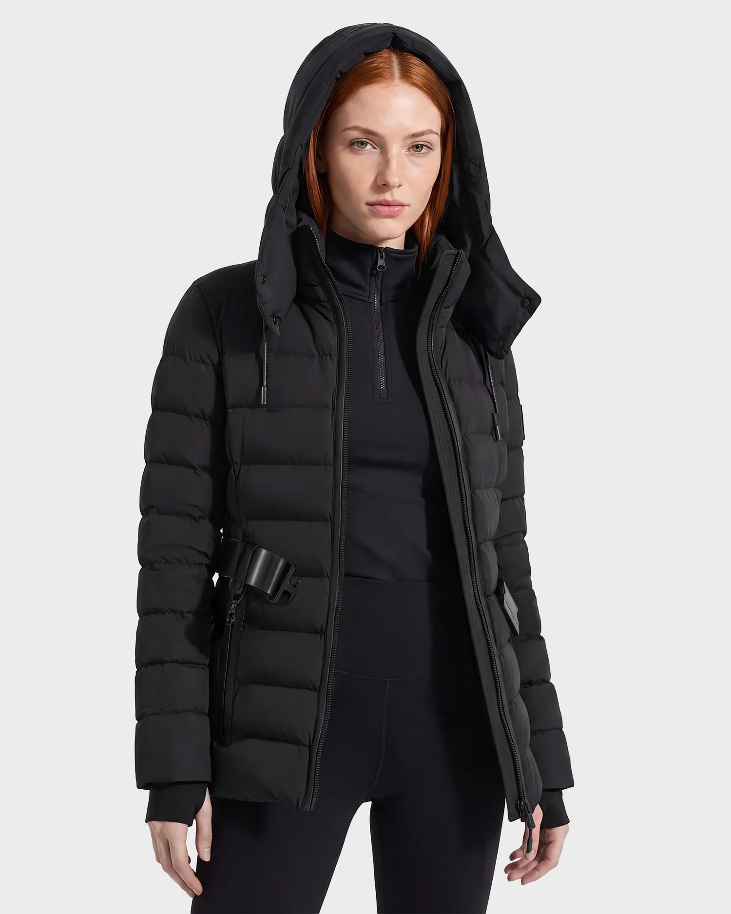 A woman with straight red hair wears the Rudsak PELIN S - 8124813 Black quilted puffer jacket and black pants, standing against a light gray background.