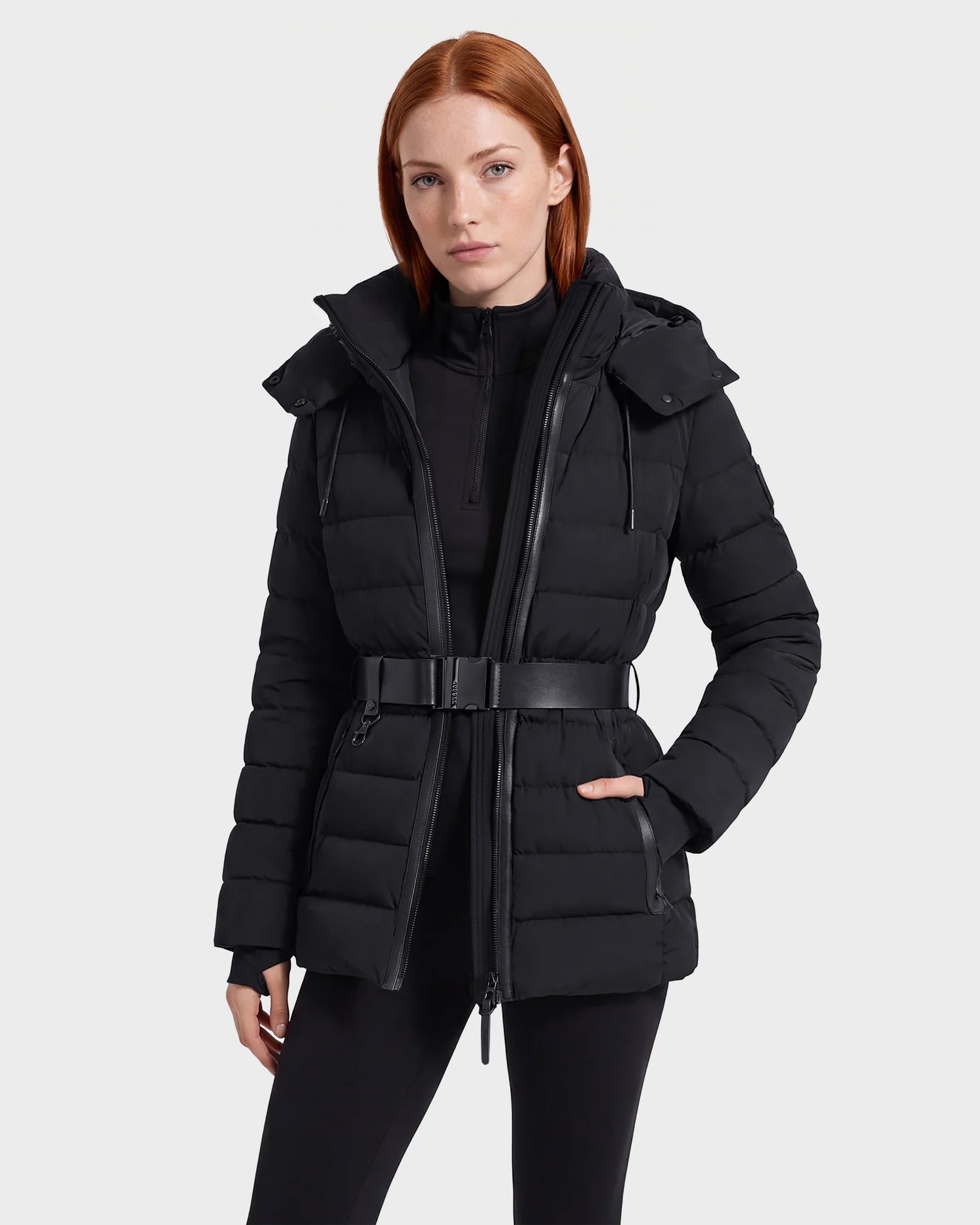 A person wearing the Rudsak PELIN S - 8124813 Black belted quilted puffer jacket over a black top and leggings stands facing forward against a plain light gray background.
