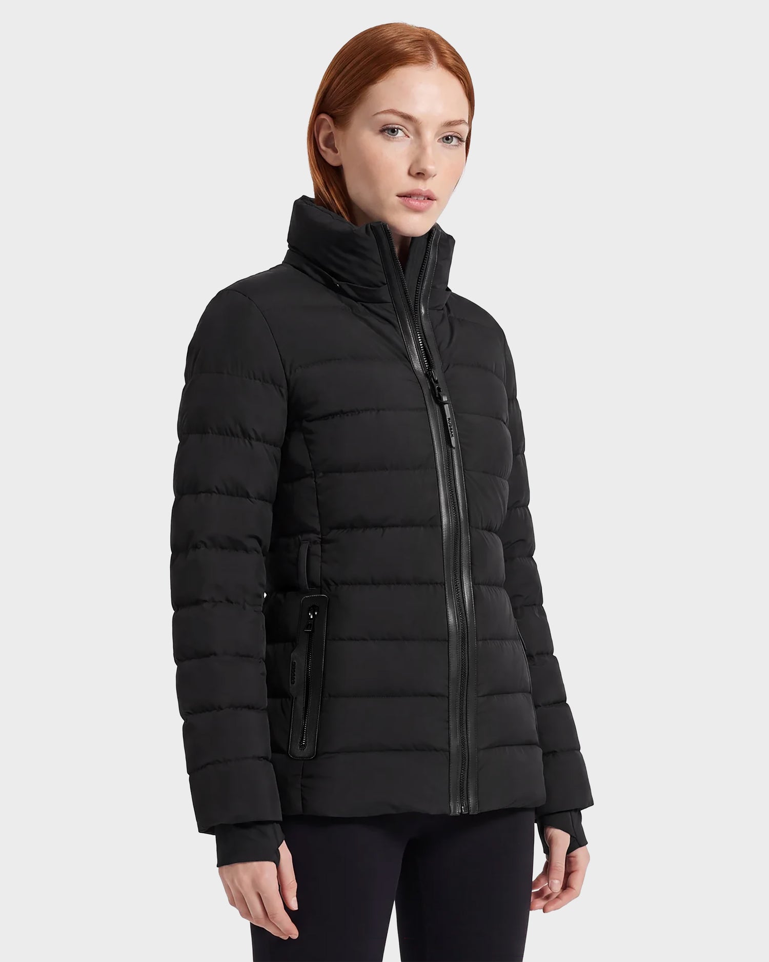 A woman with straight red hair wears the Rudsak PELIN S - 8124813 Black quilted puffer jacket, featuring a high collar and zip closure, standing against a plain light gray background.