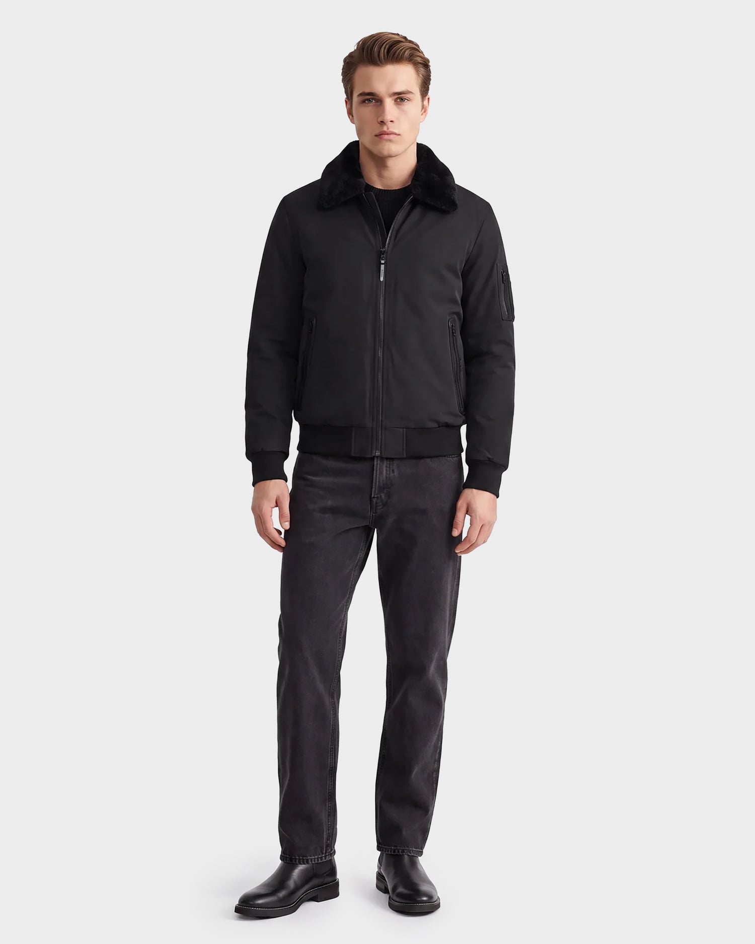 A man stands facing forward against a plain white background wearing the Rudsak REECE - 6124592 Black men's bomber jacket with a removable shearling collar, paired with black jeans and black boots.