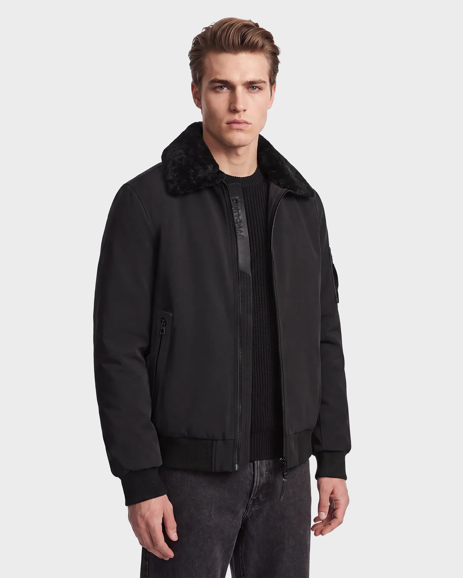 A man stands facing forward with a neutral expression against a plain light background, wearing the Rudsak REECE - 6124592 Black men's bomber jacket with a removable shearling collar, paired with a black sweater and dark pants.