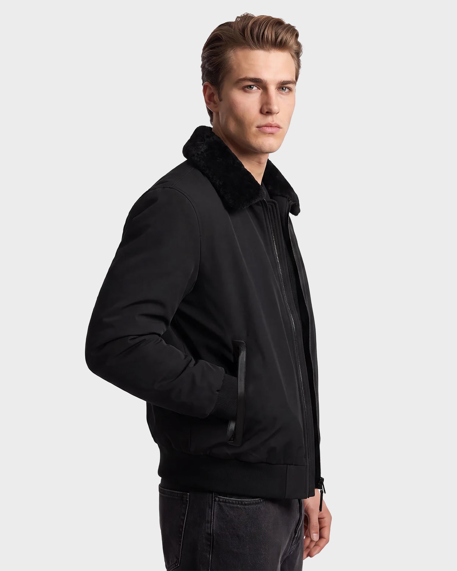 A man wearing the Rudsak REECE - 6124592 Black men's bomber jacket with a removable shearling collar stands facing slightly to the side, one hand in his pocket, against a plain light background.