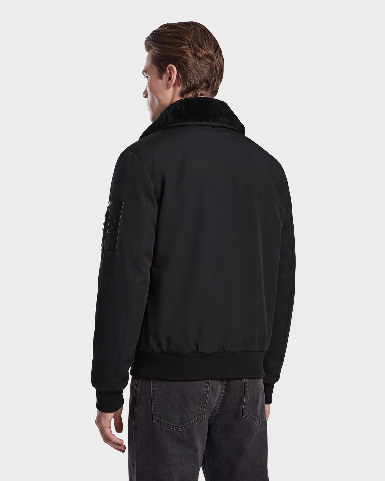 A man faces away, wearing the Rudsak REECE - 6124592 Black men's bomber jacket with a removable shearling collar and black jeans, standing against a plain white background.