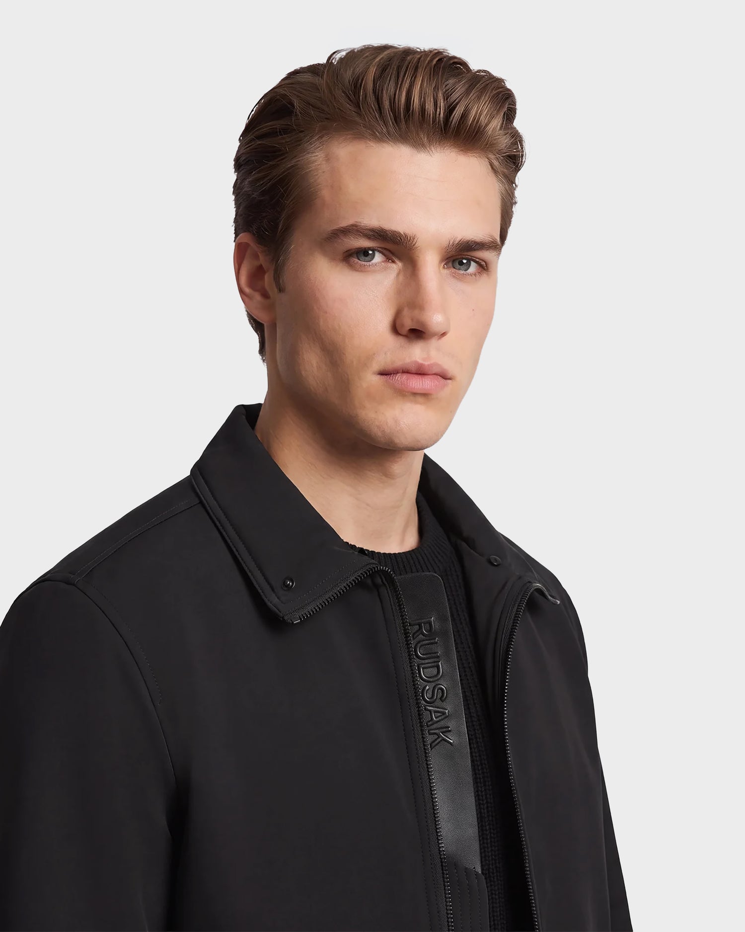A young man with short brown hair wearing the Rudsak REECE - 6124592 Black men's bomber jacket, which features eco-fill insulation and a removable shearling collar, layered over a black shirt as he looks slightly to the side against a plain light background.