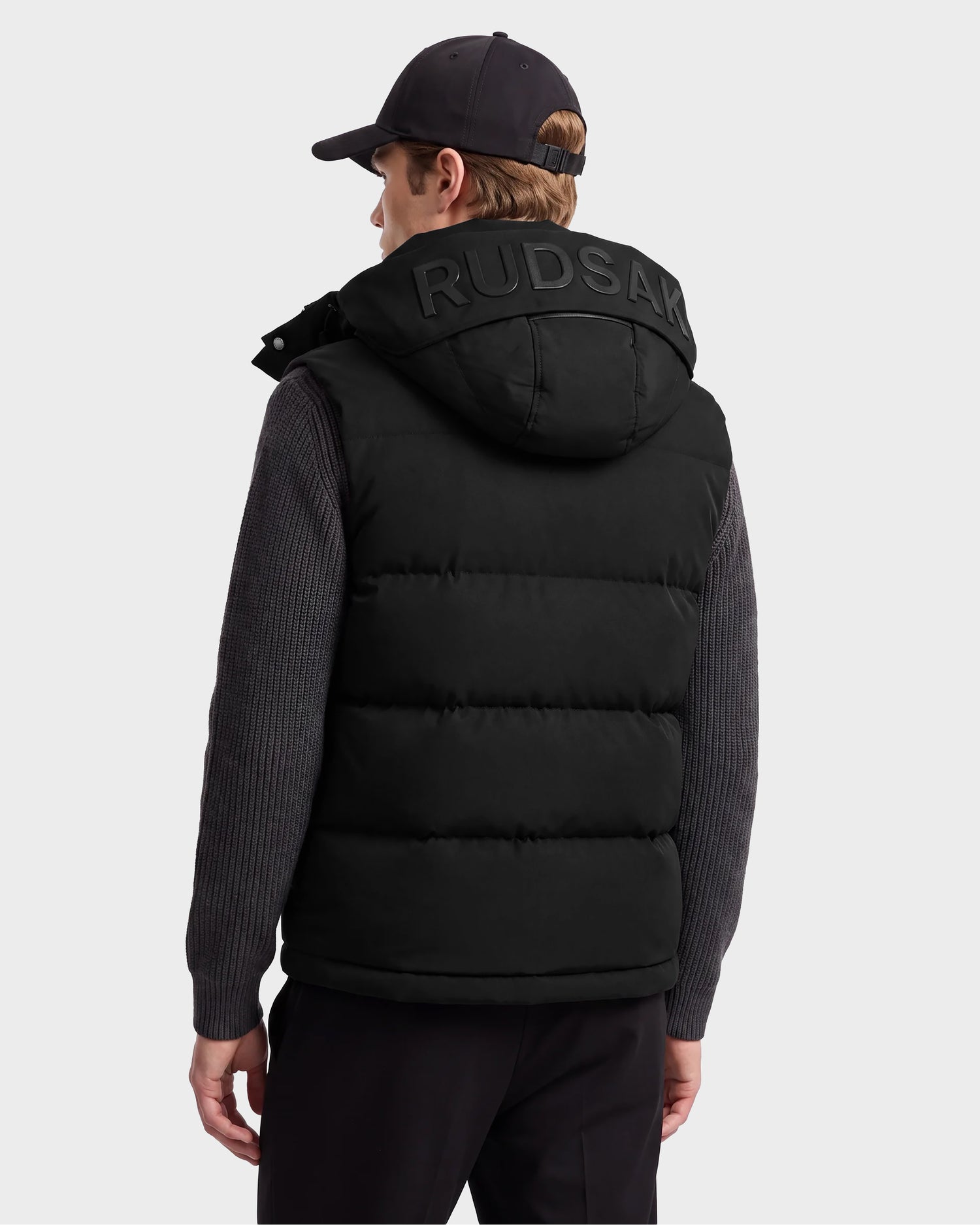 A person is seen from the back wearing the Rudsak REN M - 6125691 Black hooded puffer vest, featuring a prominent “RUDSAK” logo on the hood, along with a black cap and gray sweater.