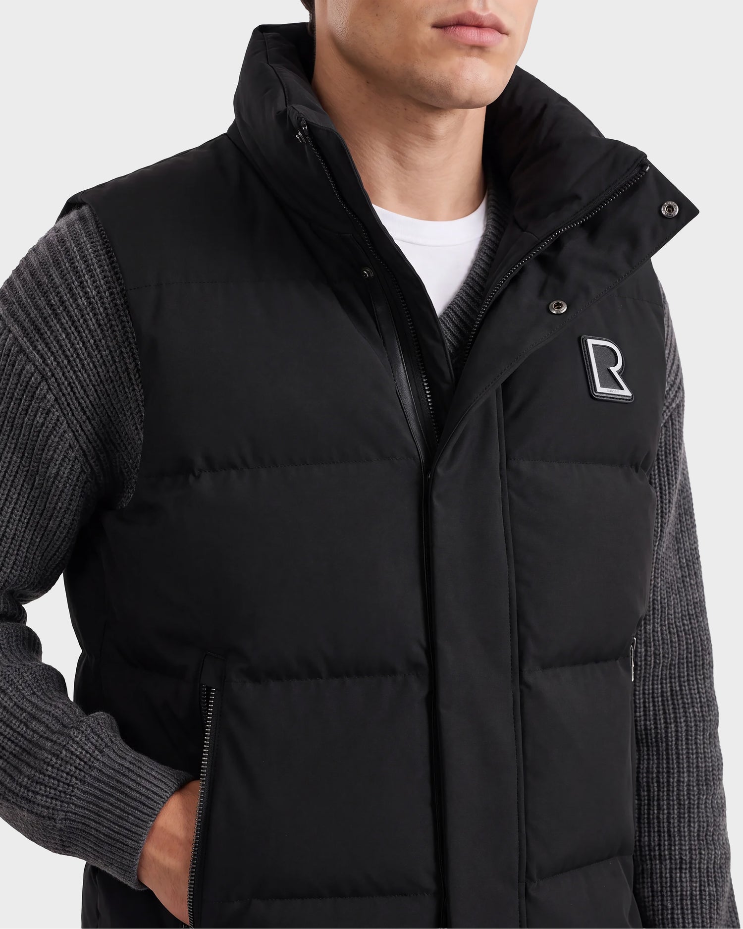 A man wearing the Rudsak REN M - 6125691 Black quilted down puffer vest with a raised collar over a gray ribbed sweater. The men’s vest features an R logo on the left chest and two zippered pockets. Only the lower half of his face and torso are visible.