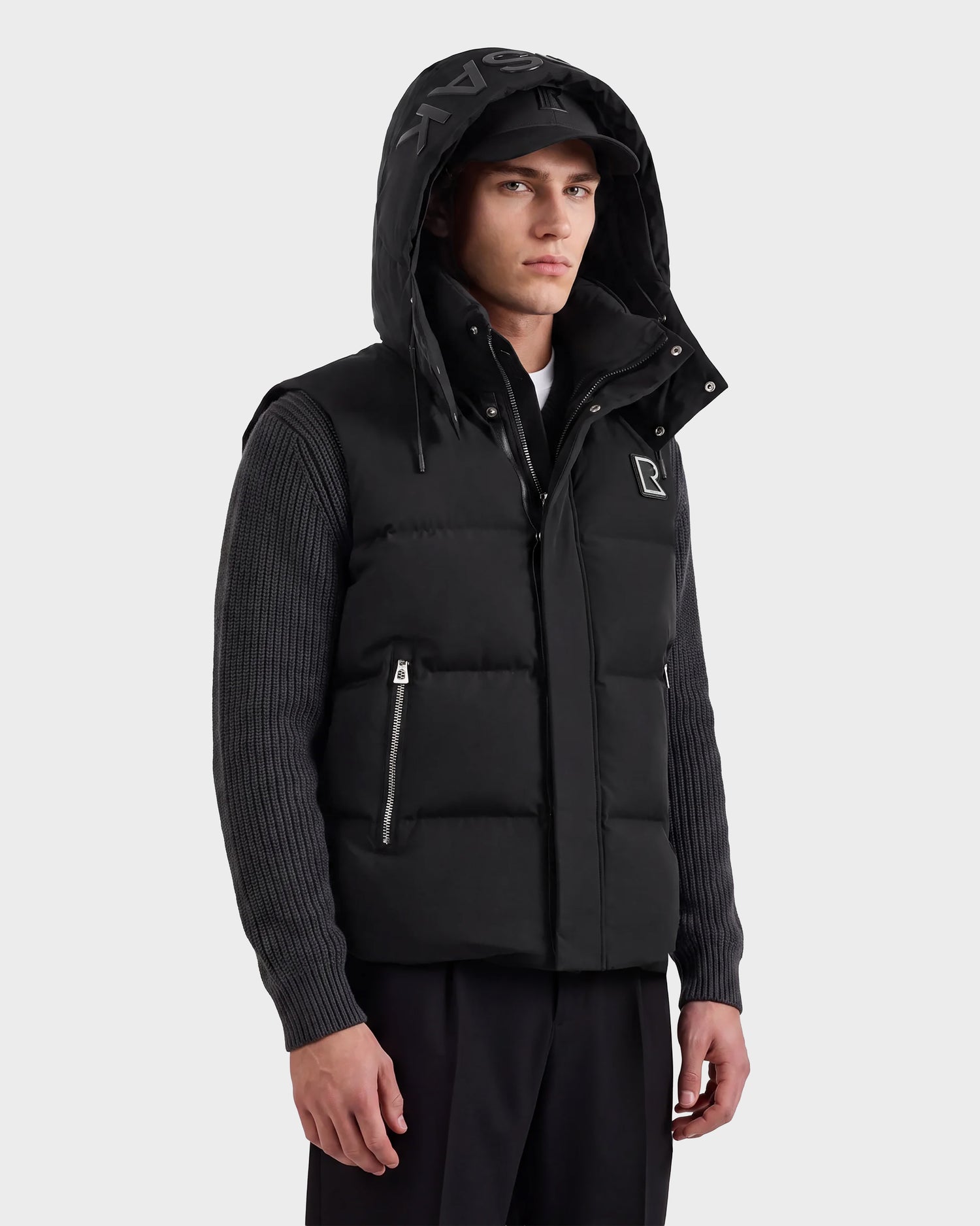 A man wearing the Rudsak REN M - 6125691 Black hooded vest, a black baseball cap, and a ribbed gray long-sleeve shirt stands against a plain light background.