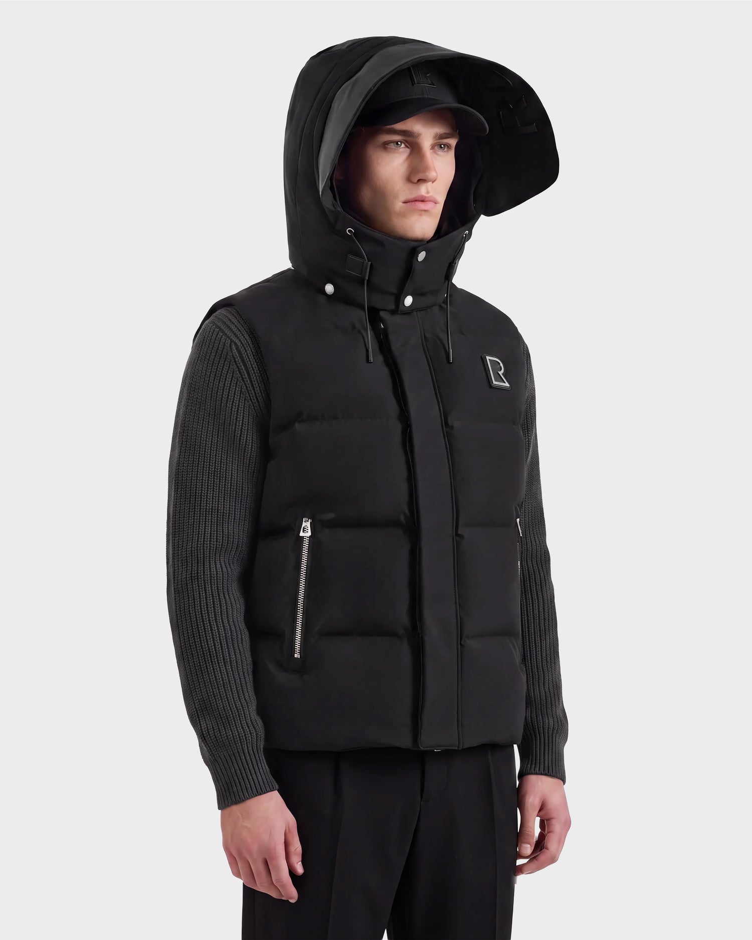 A man models the Rudsak REN M - 6125691 Black hooded puffer vest over a ribbed long-sleeve shirt and black pants, standing against a plain background and facing slightly to the side.