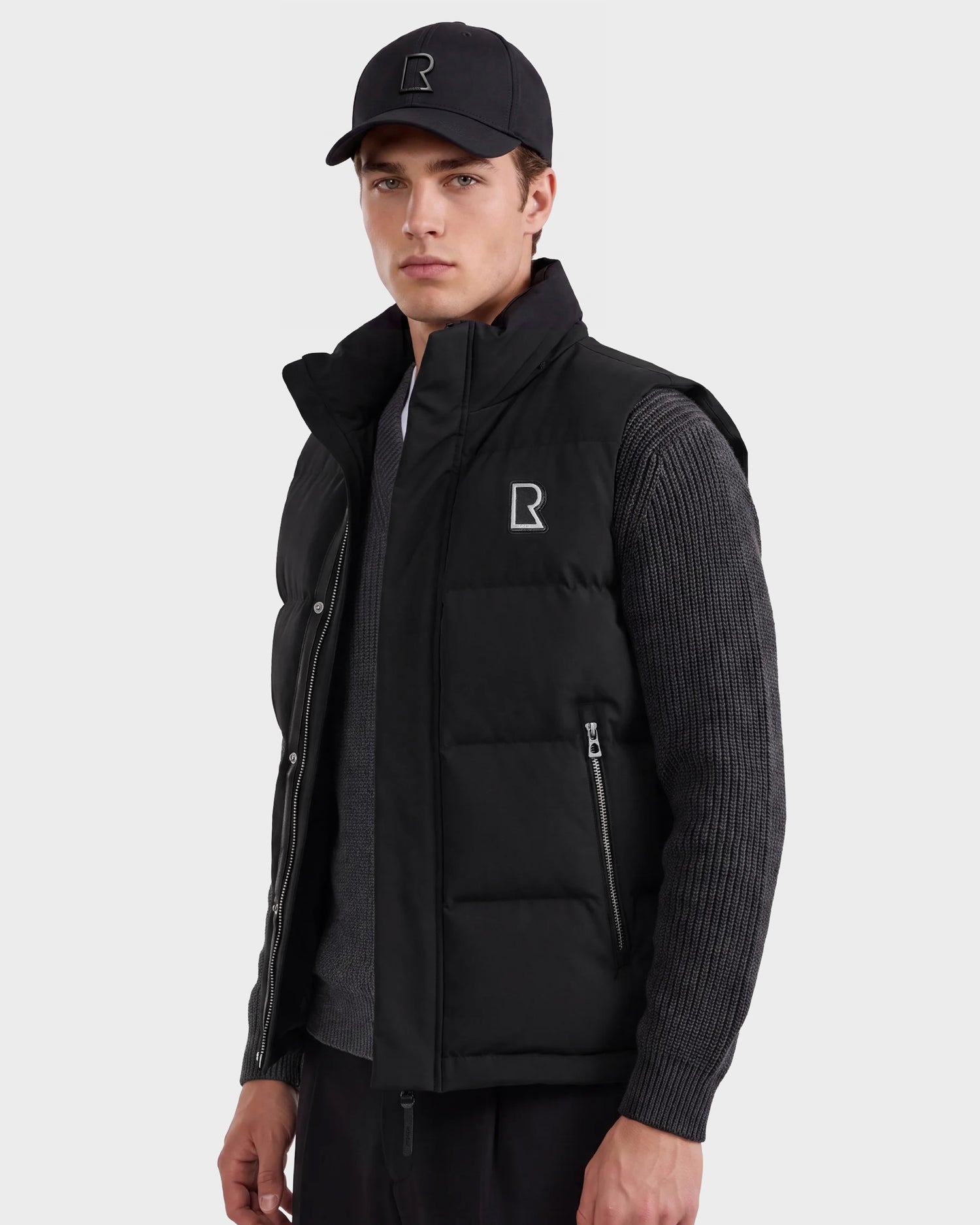 A young man wears the Rudsak REN M - 6125691 Black men’s quilted down puffer vest over a dark sweater and a black cap, each adorned with a white R logo, as he stands against a light gray background.