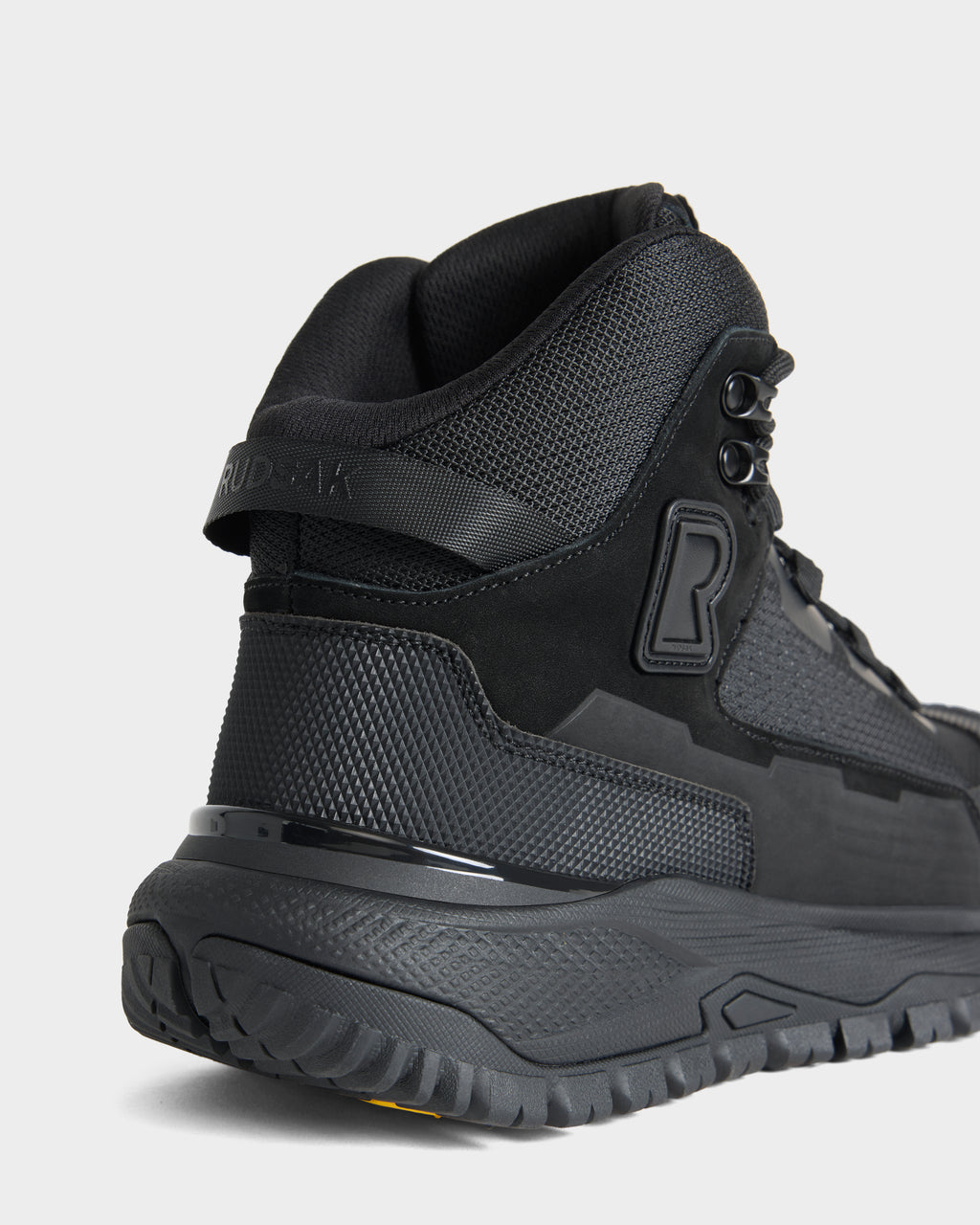 Close-up of the back and side of the REXON - 6226503 Black athletic boot by Rudsak, featuring textured details, mesh fabric, a thick sole, and a rubber “R” logo on the side. This winter-ready boot is displayed against a plain white background.