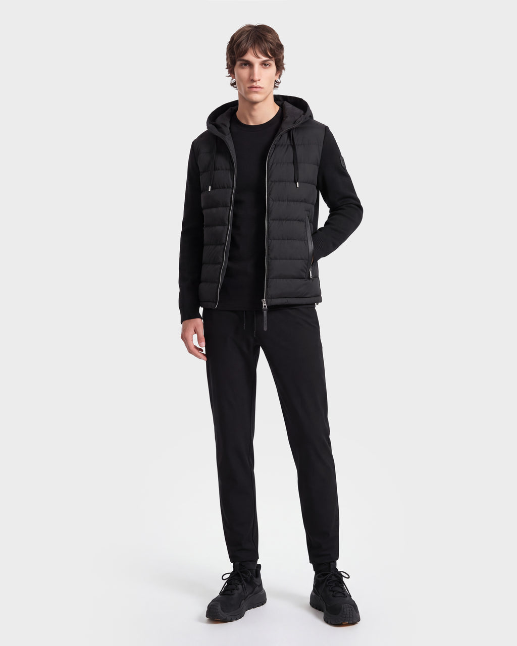 A young man stands facing forward, wearing the Rudsak RIVER HV - H125503 black hooded down jacket, paired with black pants and black shoes, against a plain white background.