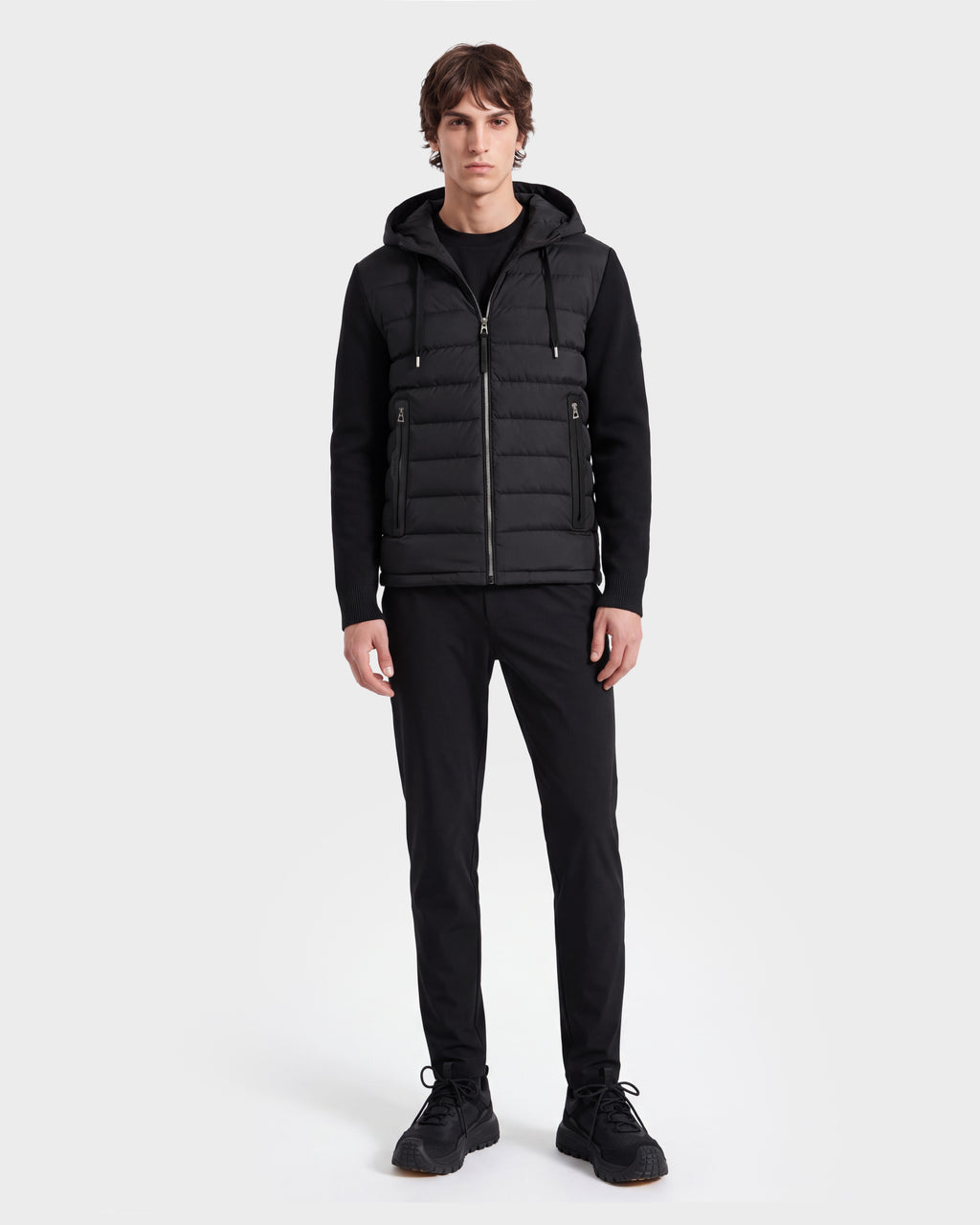 A person stands facing forward on a plain white background, wearing the Rudsak RIVER HV - H125503 Black limited edition black hooded puffer vest over a black long-sleeve shirt, paired with black pants and black sneakers.