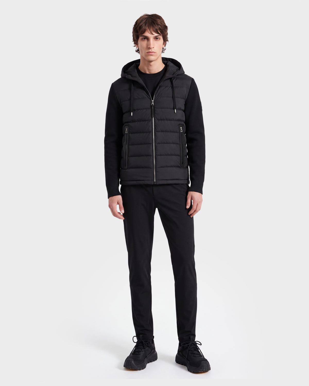 A person stands facing forward on a plain white background, wearing the Rudsak RIVER HV - H125503 Black limited edition black hooded puffer vest over a black long-sleeve shirt, paired with black pants and black sneakers.