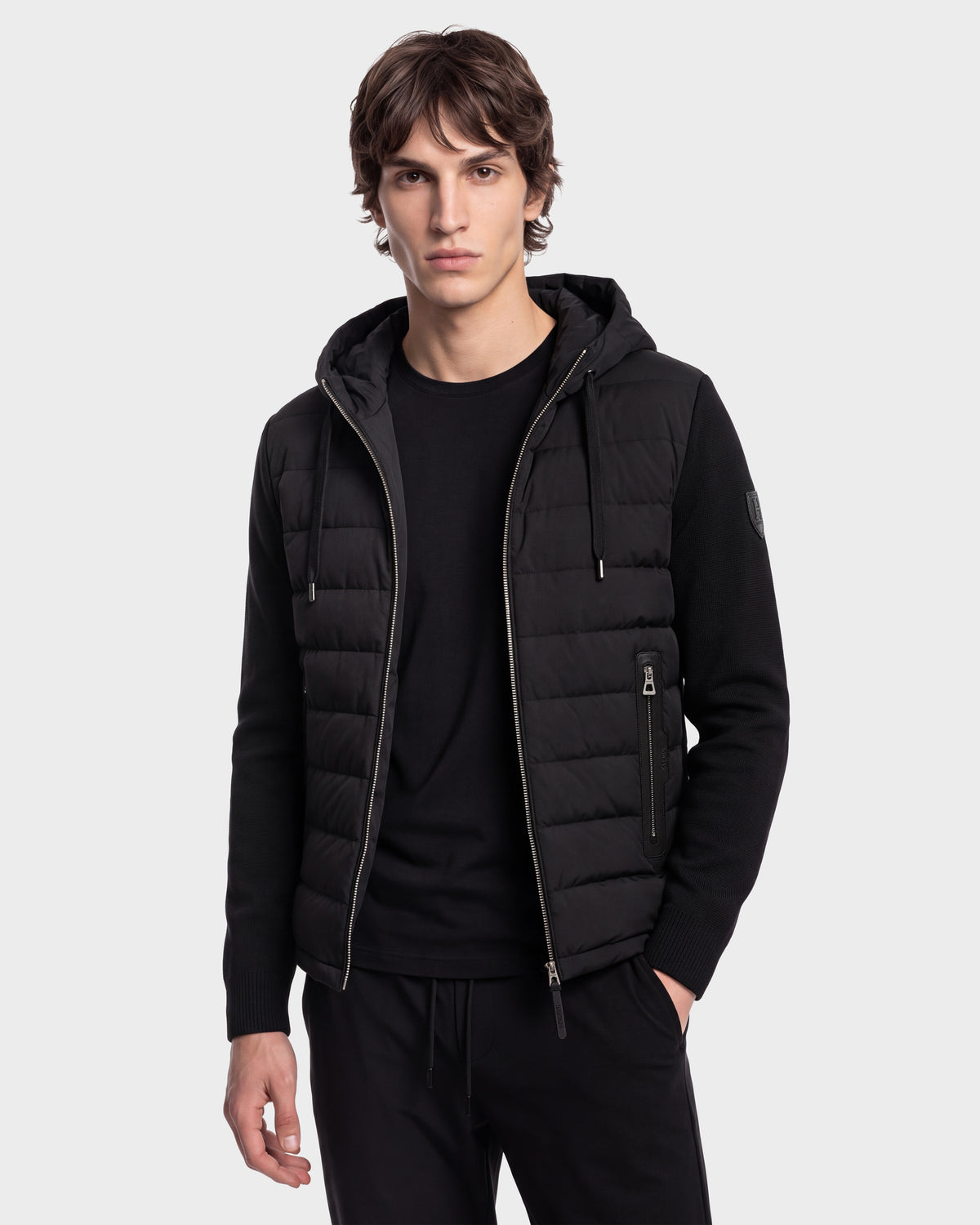 A young man with wavy brown hair stands against a plain light gray background, wearing the Rudsak RIVER HV - H125503 Black limited edition quilted hooded jacket over a black shirt and pants.