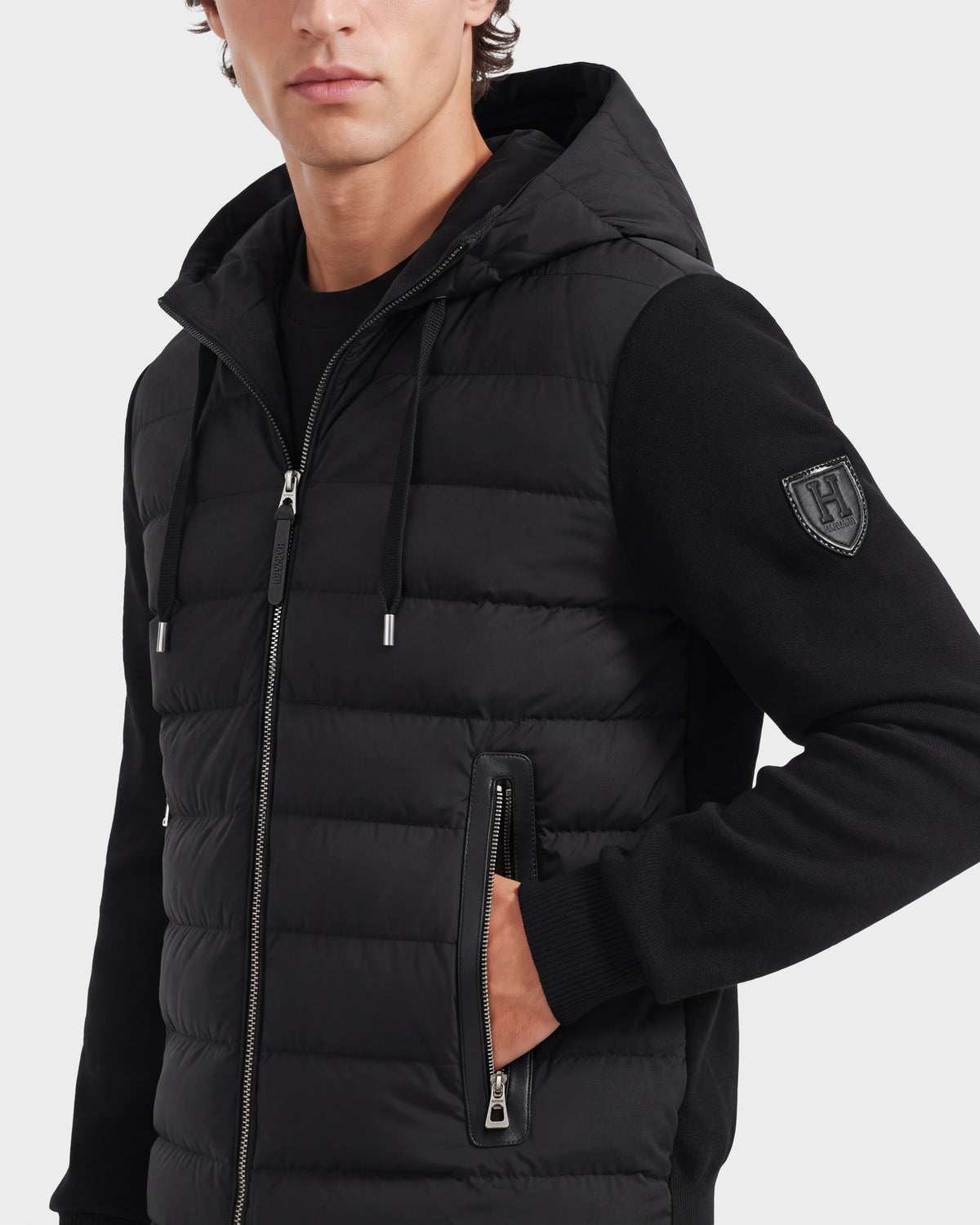 A person wearing the Rudsak RIVER HV - H125503 Black quilted down jacket with a hood, showcasing its zippered pocket and sleeve patch, poses with one hand in the pocket.