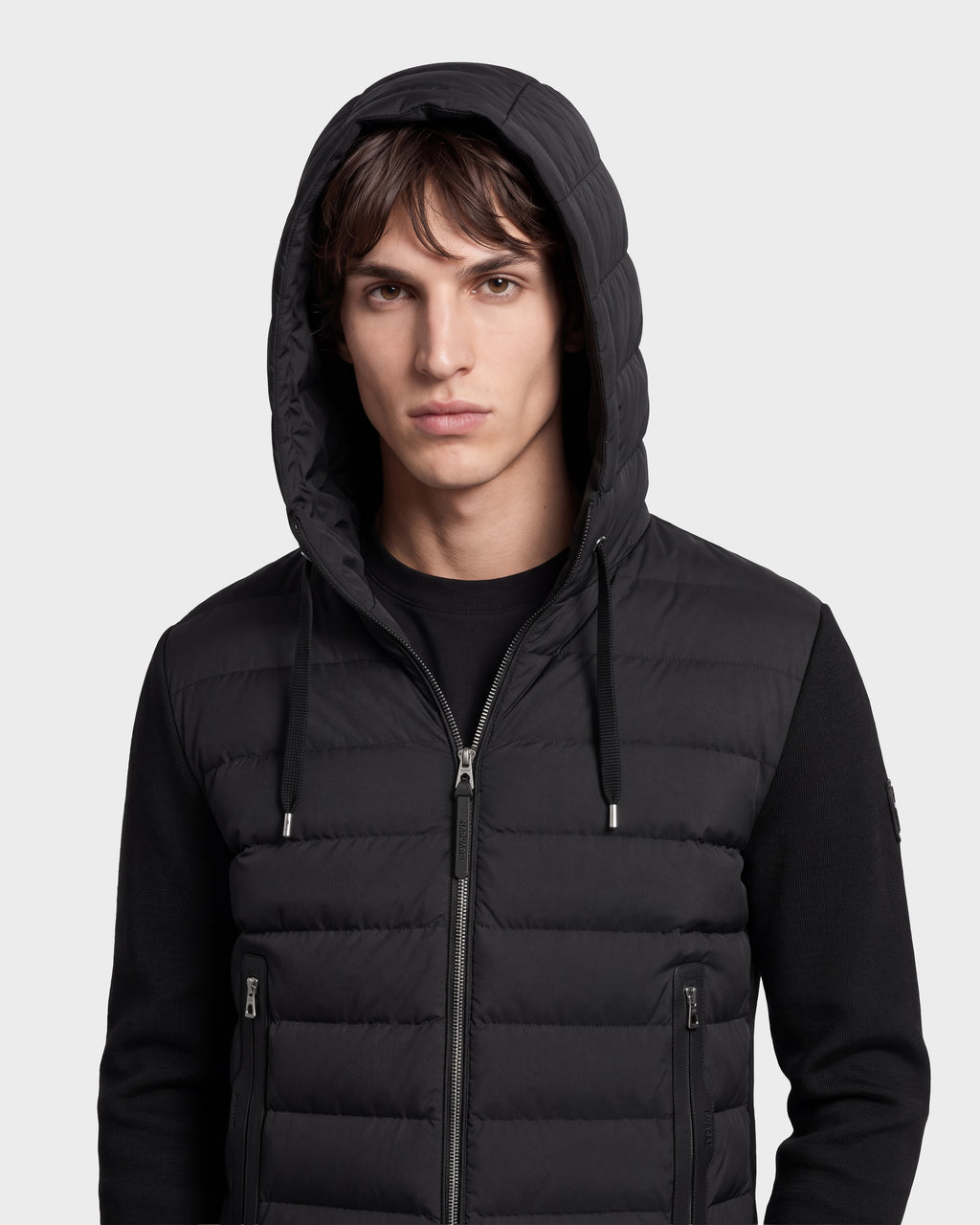 A young man stands facing forward against a plain light background, wearing the Rudsak RIVER HV - H125503 Black limited edition hooded down jacket layered over a black long-sleeve shirt.